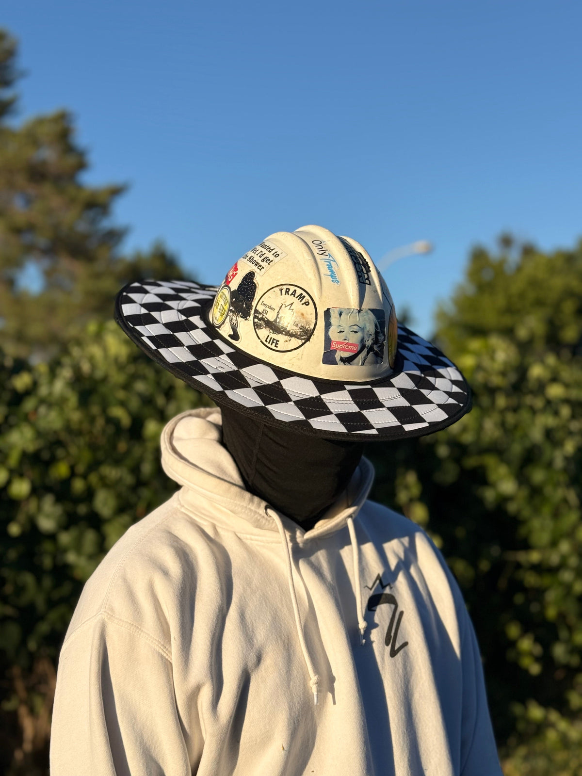 Man wearing a checkered hard hat sunshade from tramp life. Trees in the background and blue skies with a white sweatshirt 