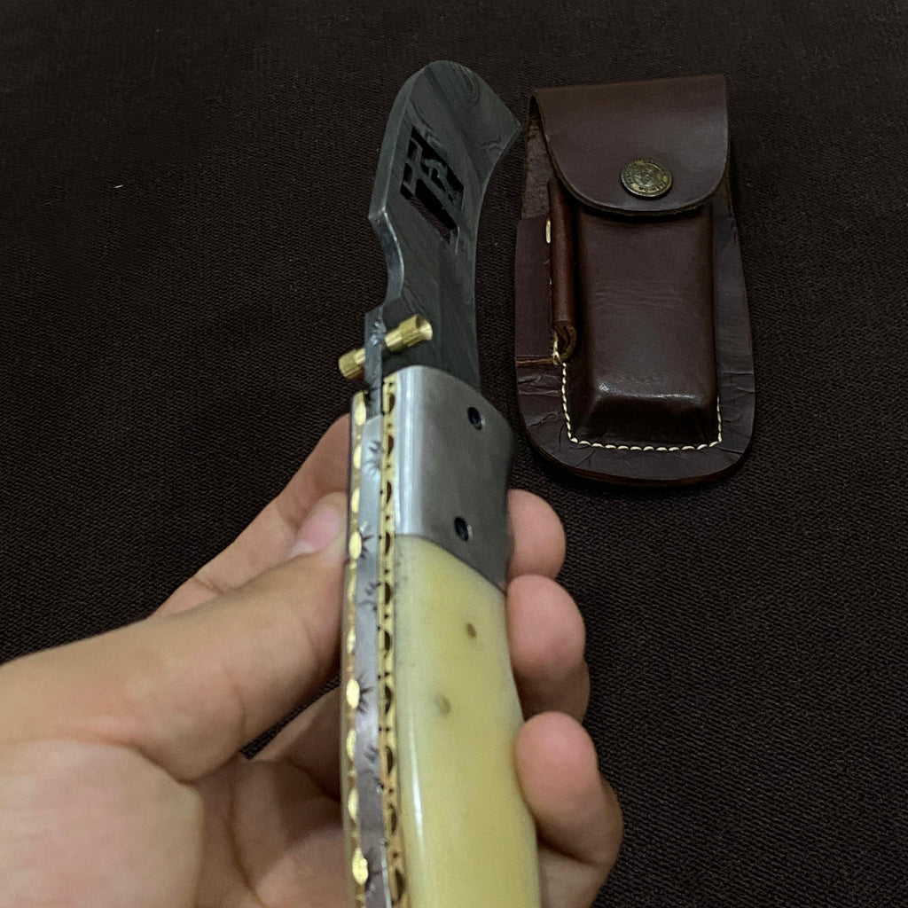 Hand holding a folding knife with a decorative handle next to a leather sheath on a dark background