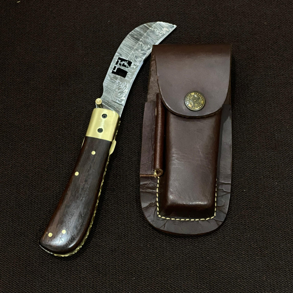 foldable damascus steel hawksbill knife with wood handle next to dark leather sheath