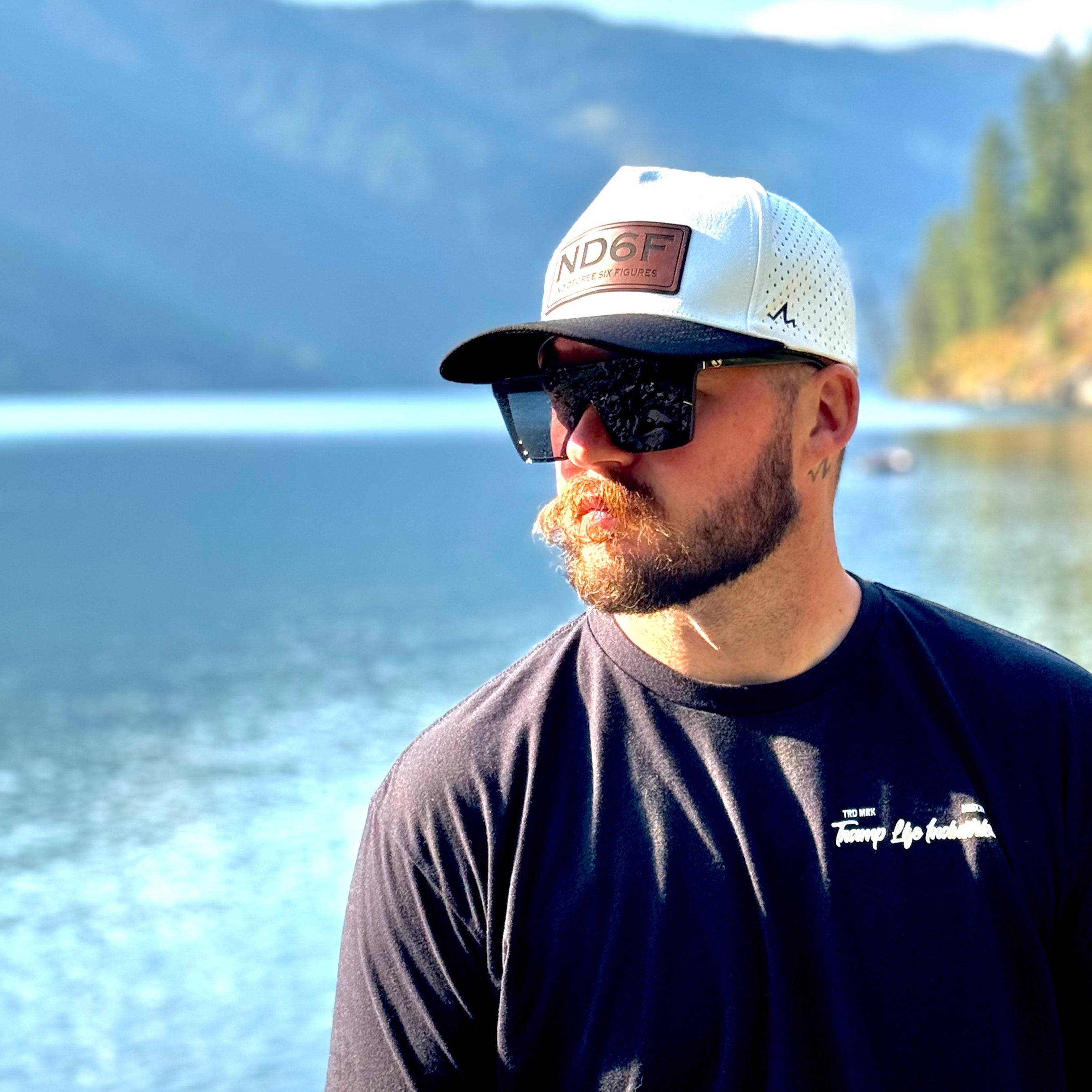 Man wearing a cap and sunglasses with a scenic lake and mountains in the background