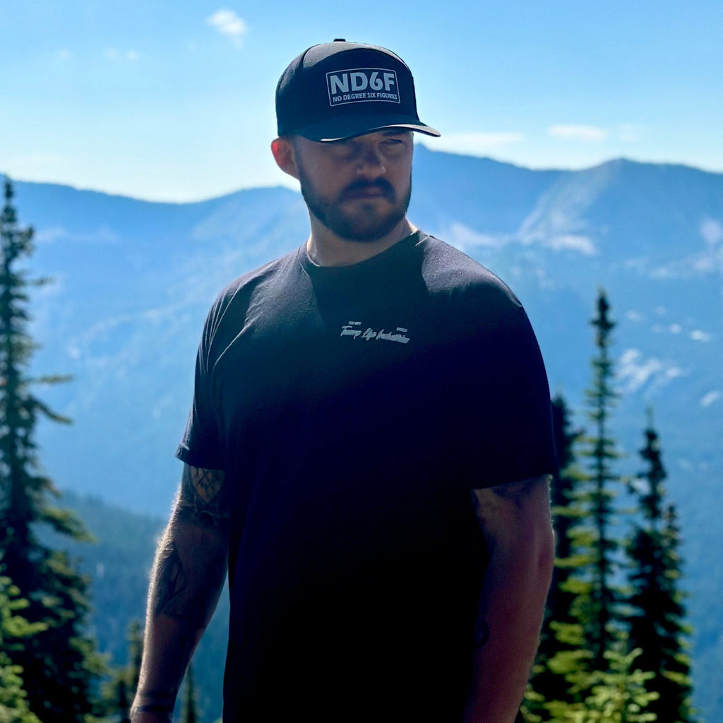 man standing in front of trees and mountains with the front of the stay golden shirt from tramp life showing
