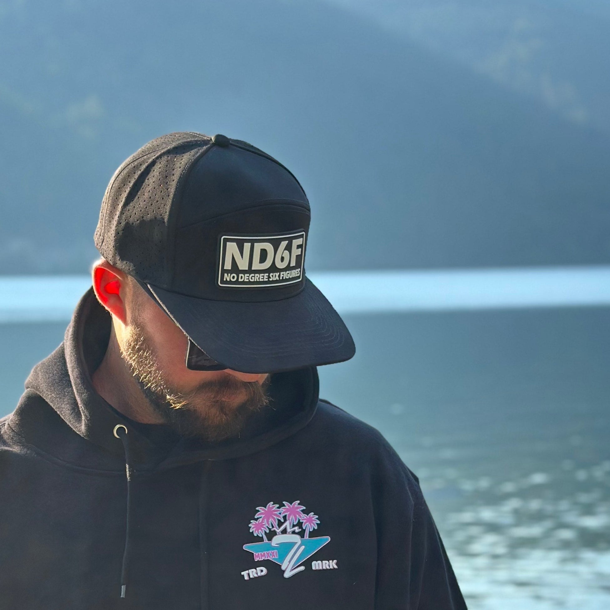 Man wearing a black cap and hoodie with a logo, standing by a lake with mountains in the background. 