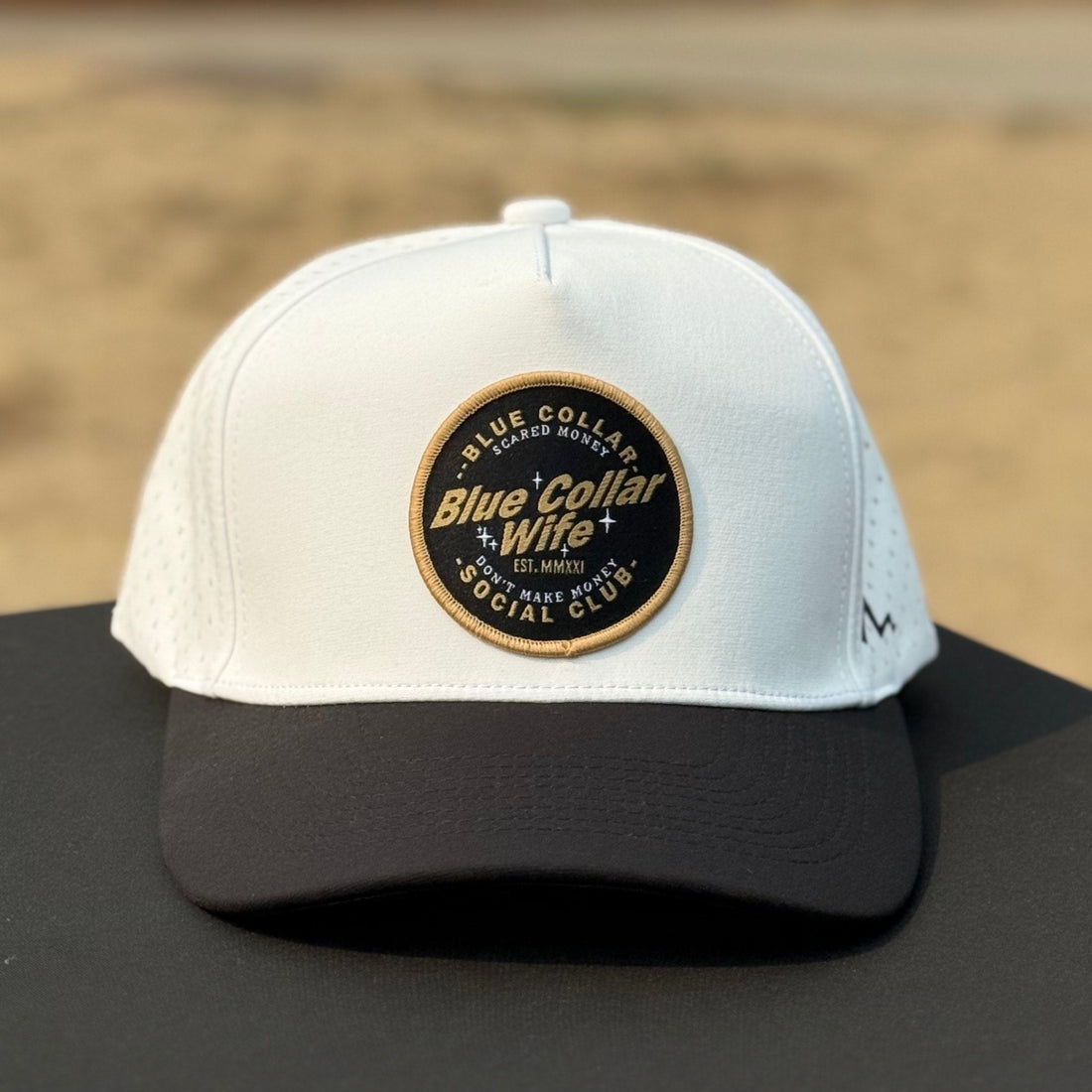 White cap with a logo on a black surface outdoors. hat patch says blue collar wife social club