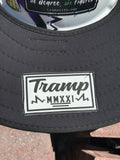 a close up of An all-black sunshade from tramp life industries with a PVC patch sewn into it with the words TRAMP on a white hard hat with tramp life stickers