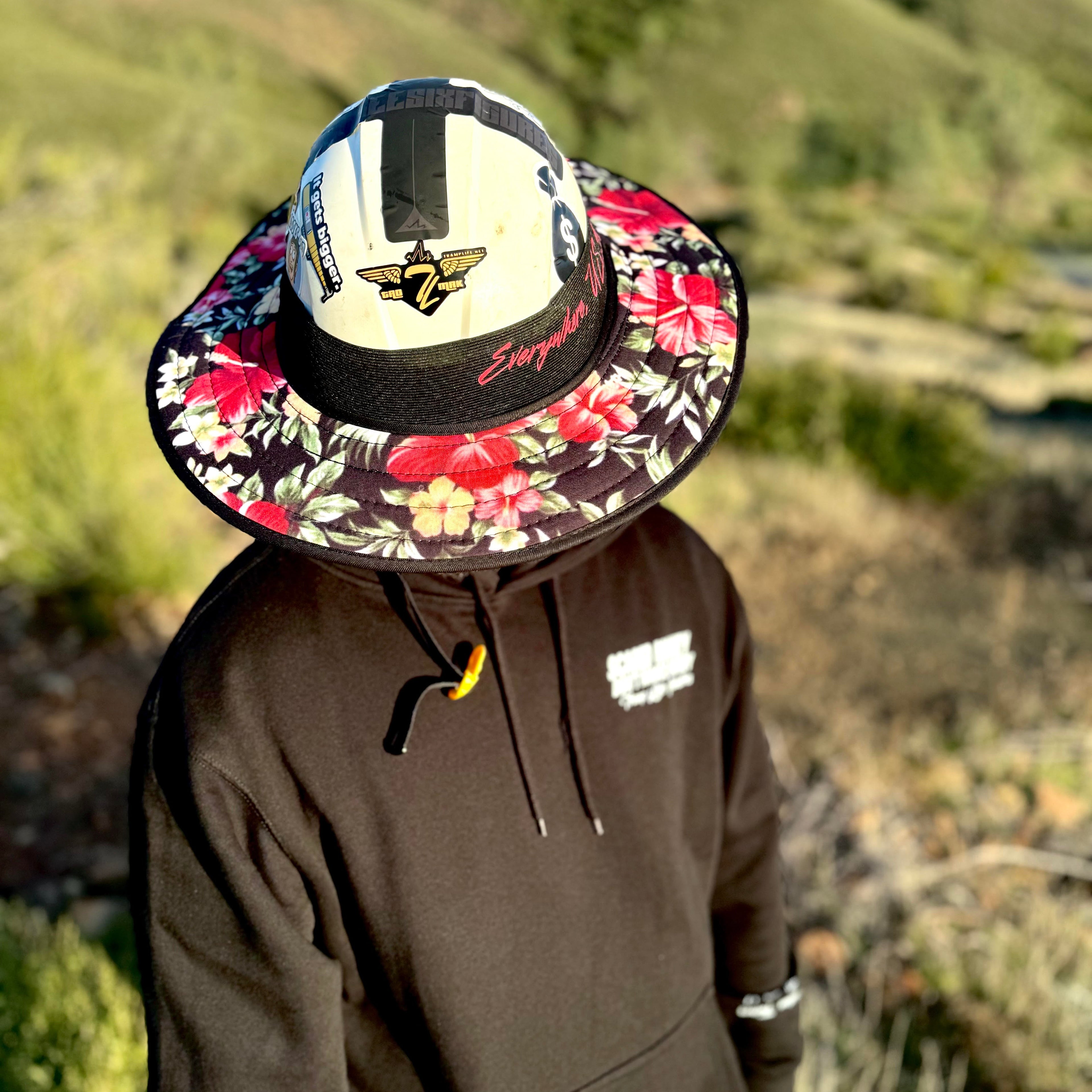 man standing on a mountain with grass in the background, he is wearing an aloha fly shade from tramp life industries. It has pink floral hibiscus flowers as a design