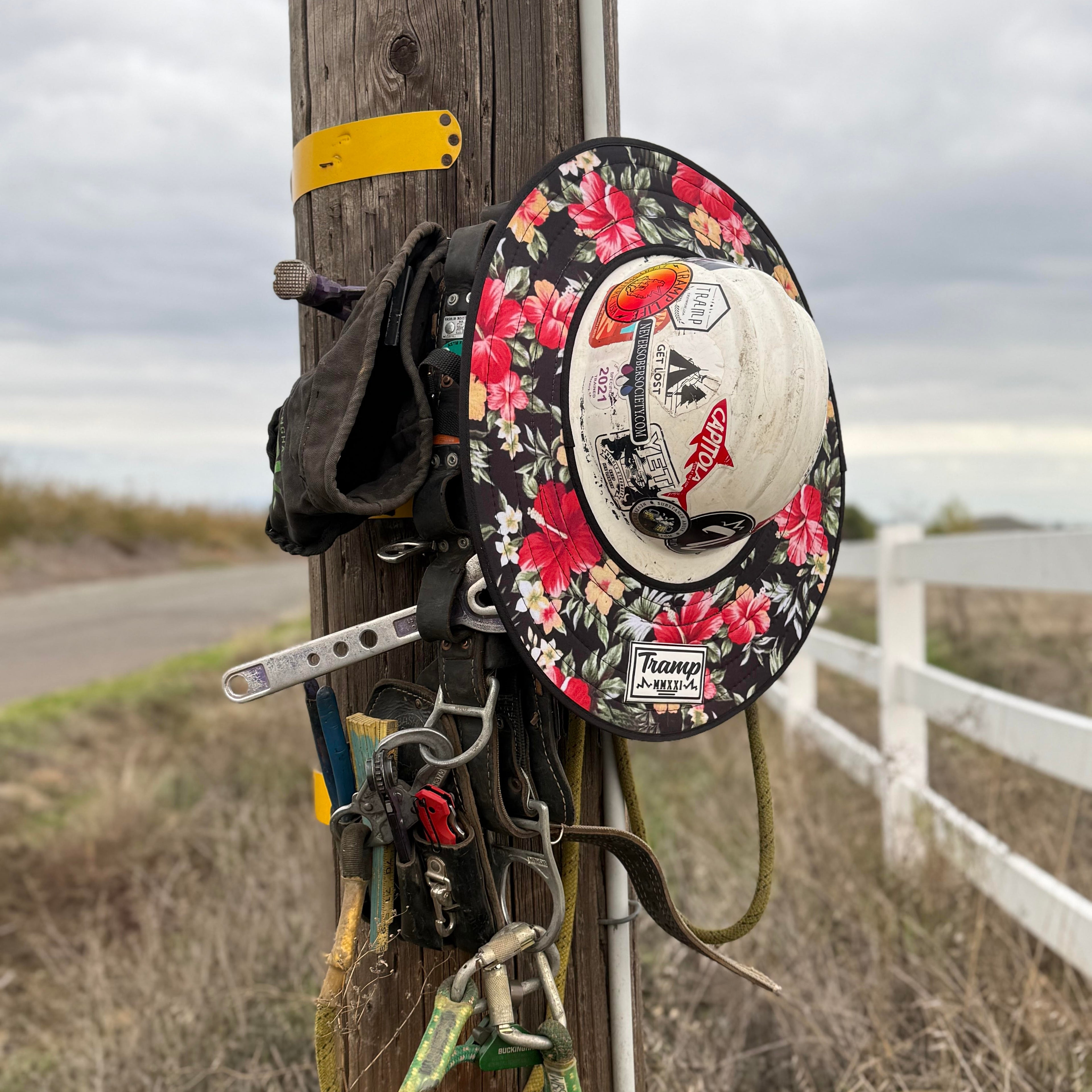 A floral print hard hat sunshade from tramp life industries. Features a Hawaiian flower print and attaches to full brim hard hats with an elastic band. the hardhat and sunshade are on a power pole hanging next to lineman climbing gear.