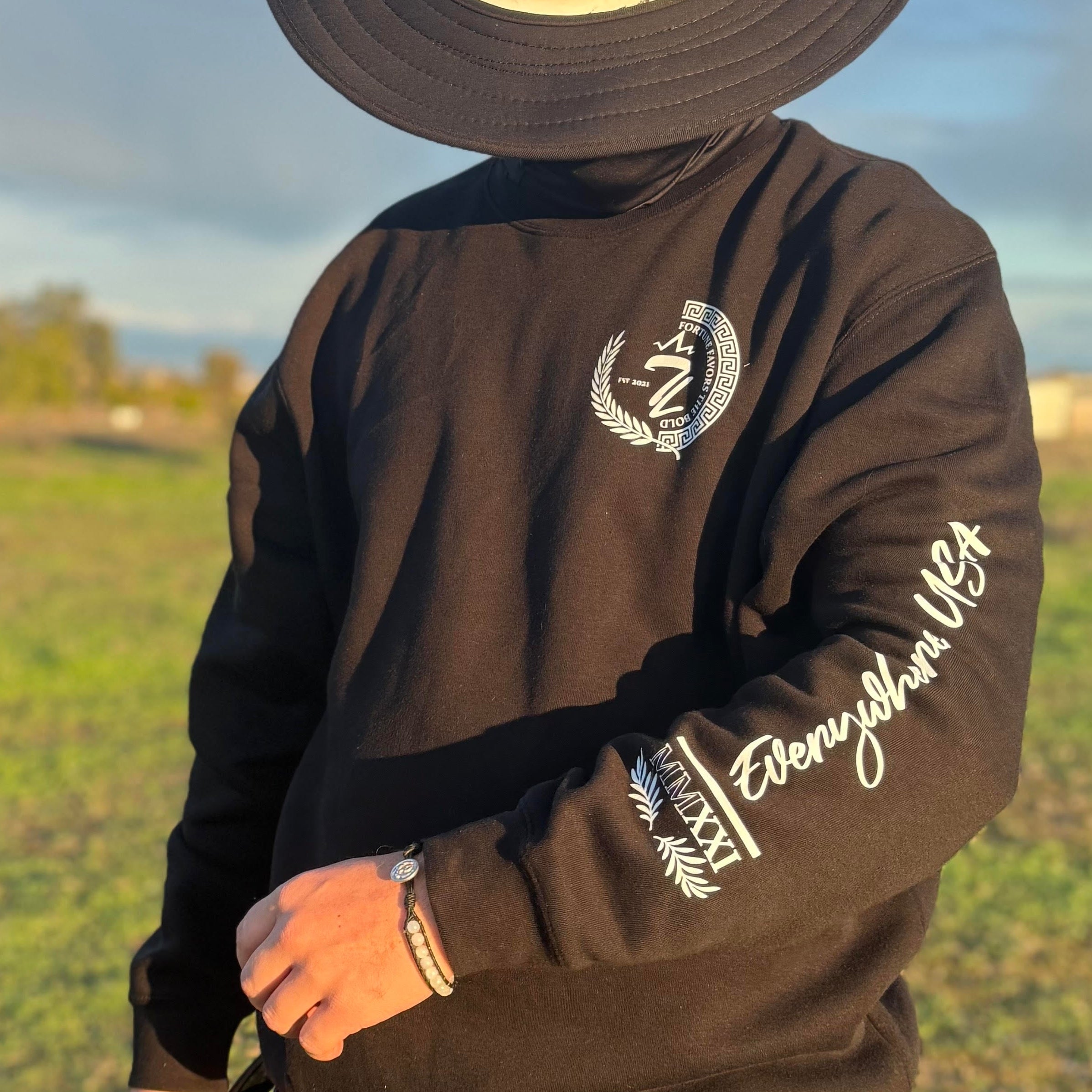 Person wearing a black sweatshirt with text and graphics, standing in an open field with a blue sky. the text says "everywhere, USA" which is a signature line from tramp life industries. It also has the numbers MMXXI
