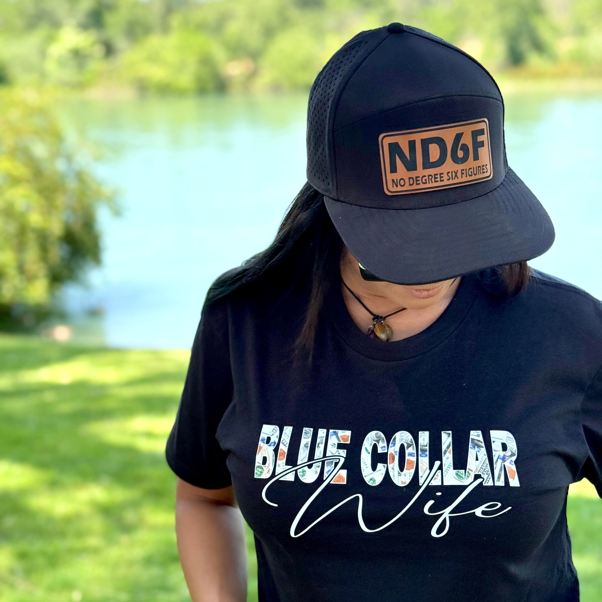 Person wearing a black cap and 'Blue Collar Wife' t-shirt outdoors by a lake. Blue Collar Wife, Blue collar woman, blue collar, ND6F. no degree, six figures 