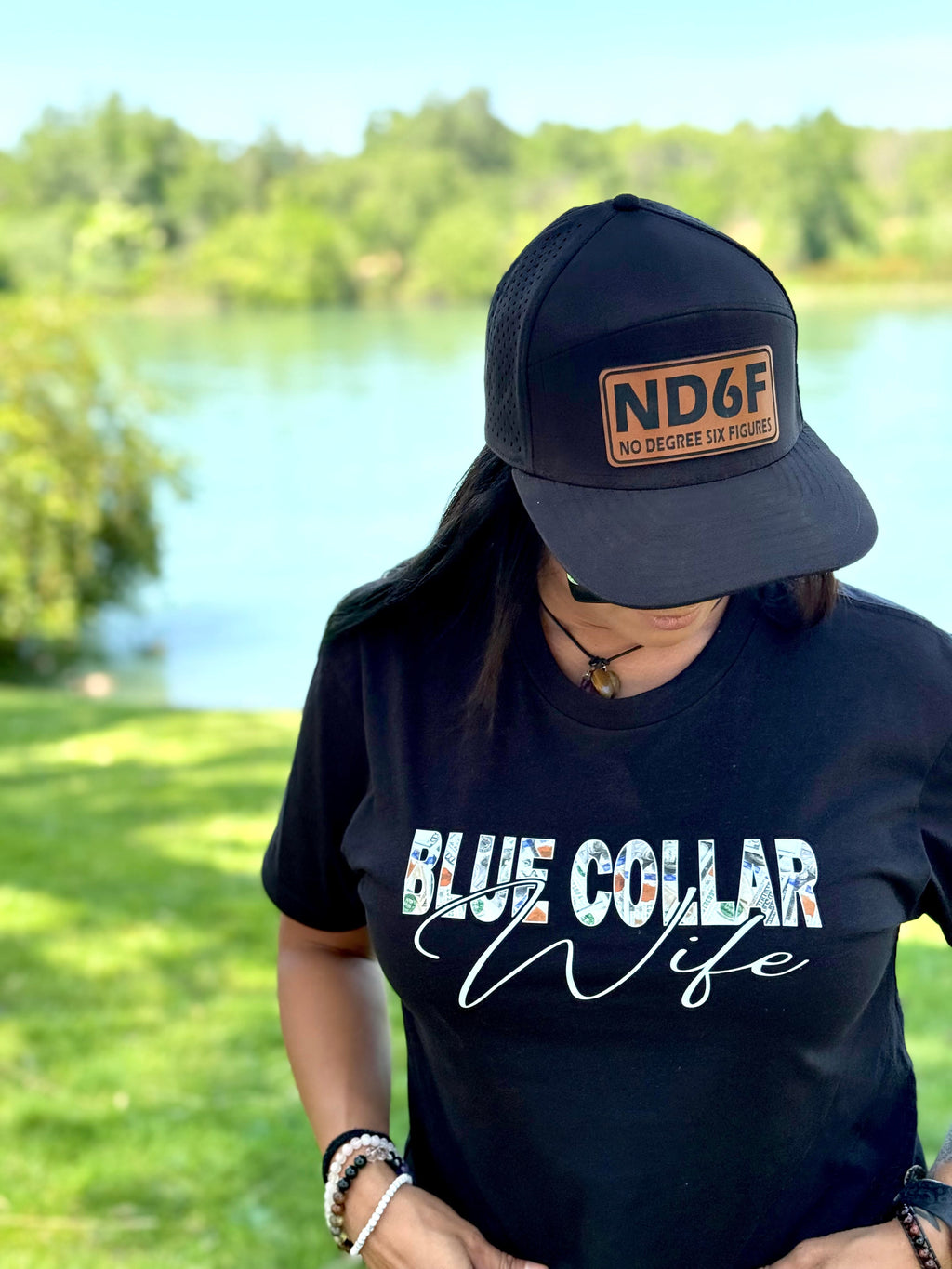 Person wearing a black cap and 'Blue Collar Wife' t-shirt outdoors by a lake. Blue Collar Wife, Blue collar woman, blue collar, ND6F. no degree, six figures 