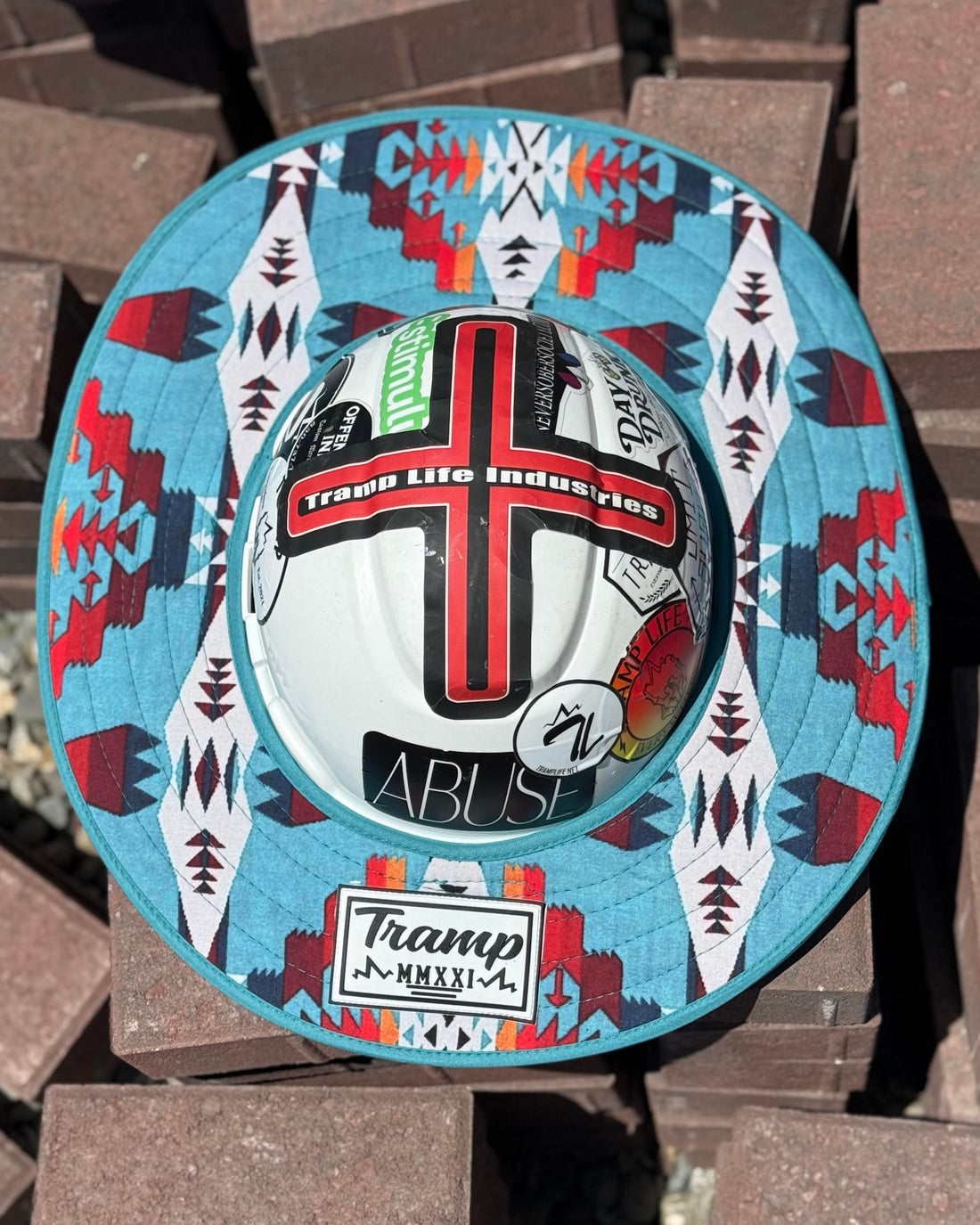 a turquoise sunshade with red, yellow, white and navy accents from tramp life industries on a white hard hat with tramp life stickers sitting on a pile of bricks