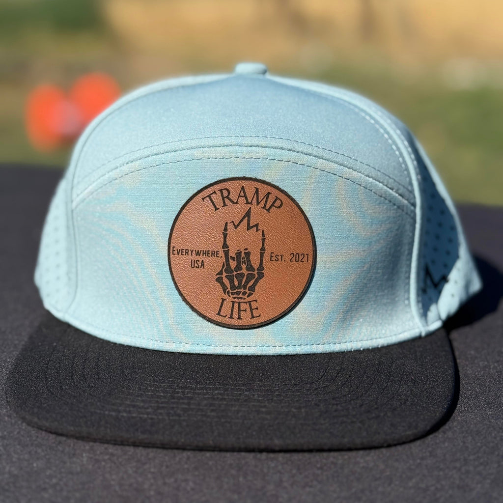 Light blue cap with brown leather patch featuring 'Tramp Life' logo on a blurred outdoor background.