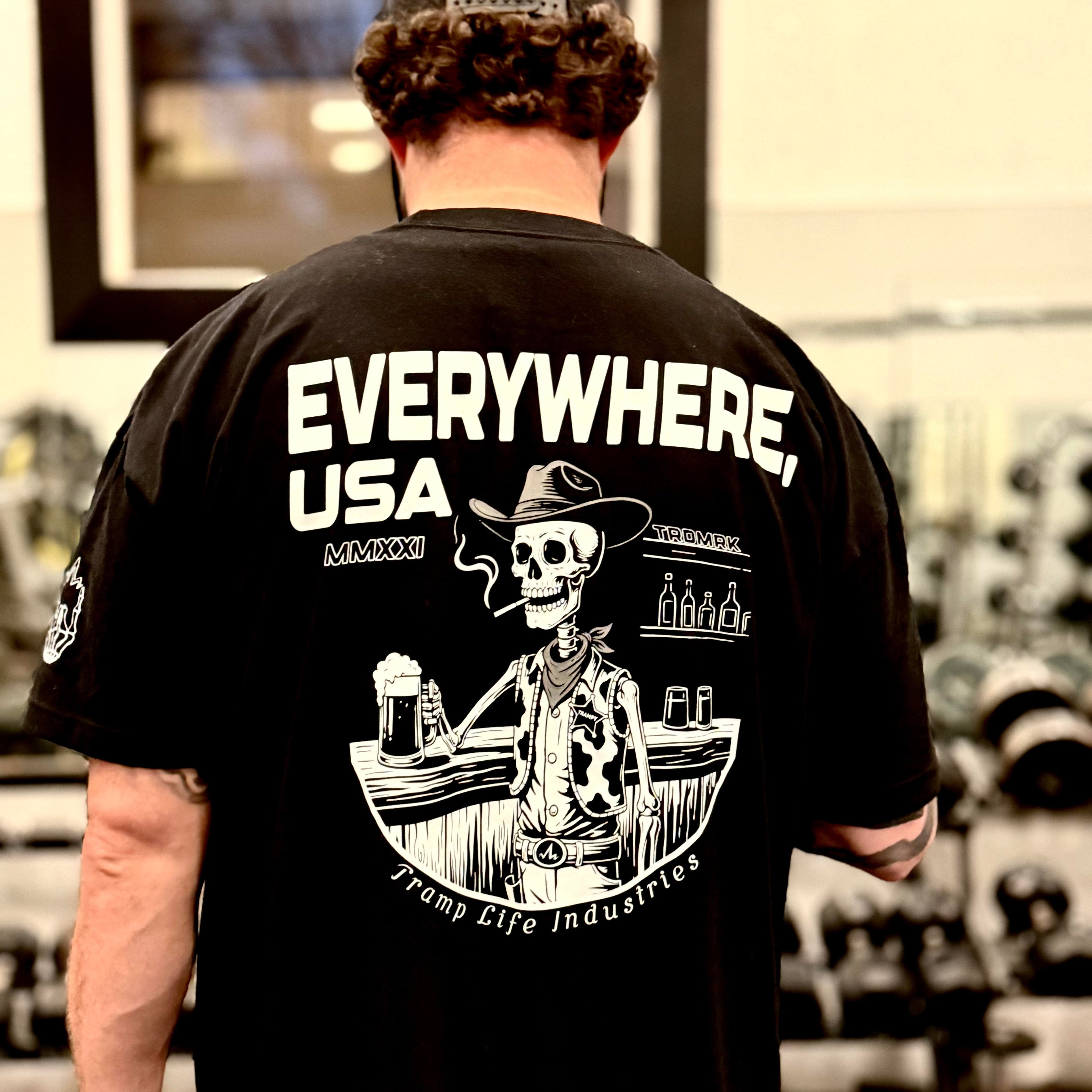 man wearing a black shirt with a skeleton on it that says everywhere usa by tramp life industries. in the gym 