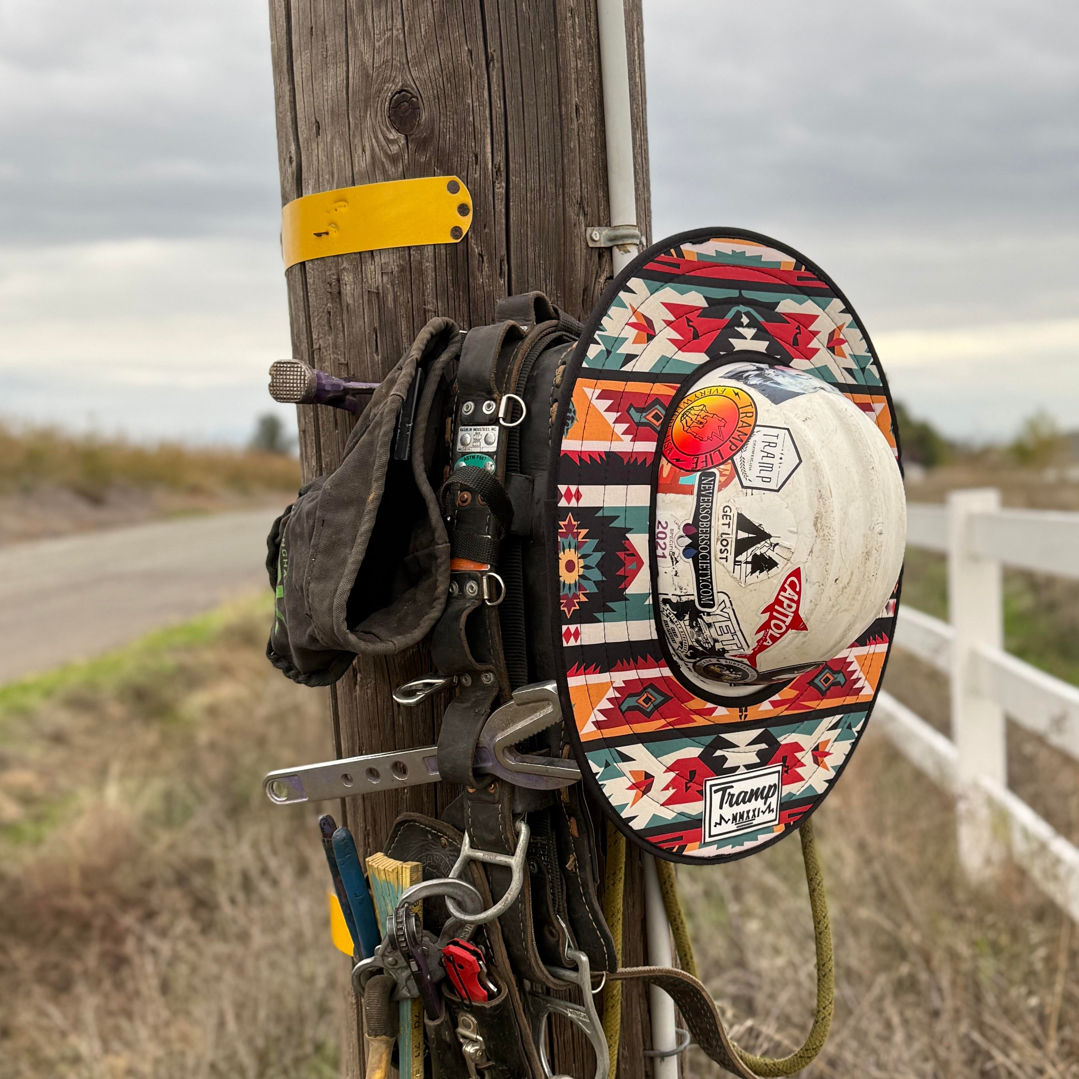 A red pendleton print hard hat sunshade from tramp life industries. Features a pendleton print with red, orange, teal, beige, nlack and yellow multicolor print and attaches to full brim hard hats with an elastic band. the hardhat and sunshade are on a power pole hanging next to lineman climbing gear.