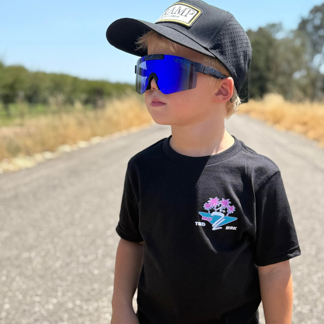 Child wearing a black t-shirt from tramp life with pink and blue palm tree and logos that says fast hands easy money, standing on a road.