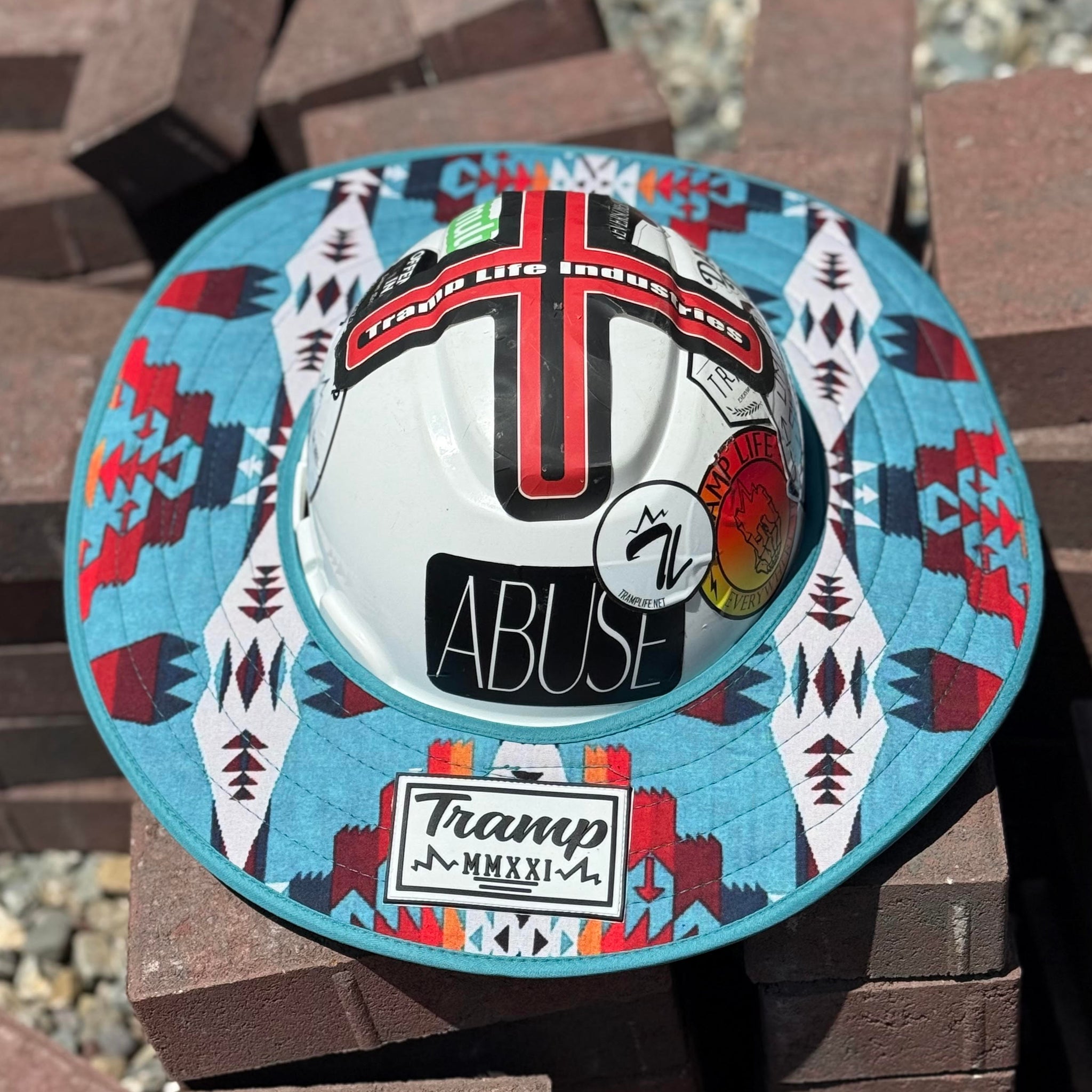 a turquoise sunshade with red, yellow, white and navy accents from tramp life industries on a white hard hat with tramp life stickers sitting on a pile of bricks