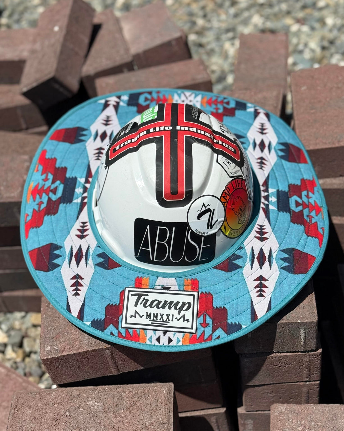 a turquoise sunshade with red, yellow, white and navy accents from tramp life industries on a white hard hat with tramp life stickers sitting on a pile of bricks