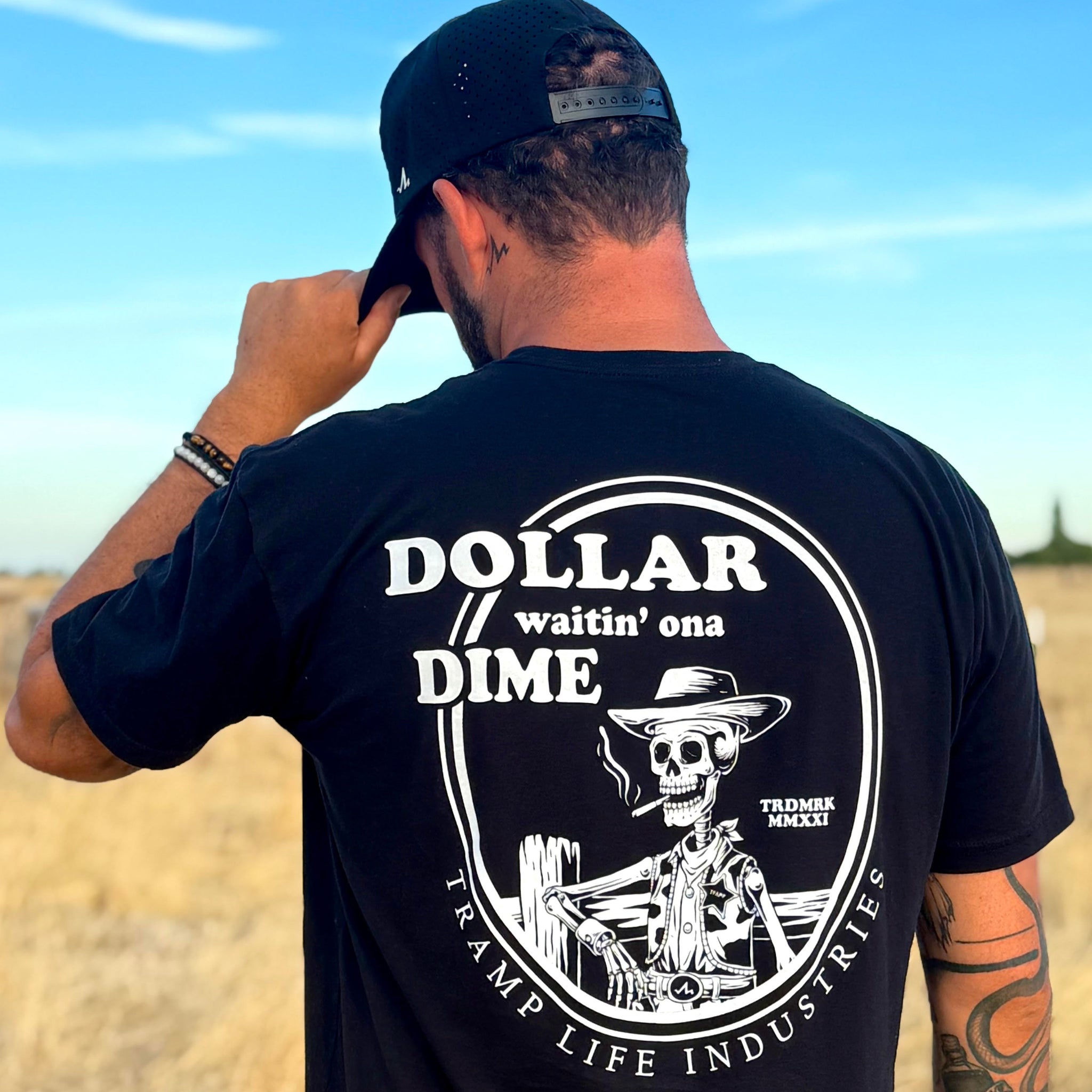 man standing in a field wearing a black tramp life shirt with the words dollar waitin' ona dime and a skeleton smoking a cigarette leaning on a fence