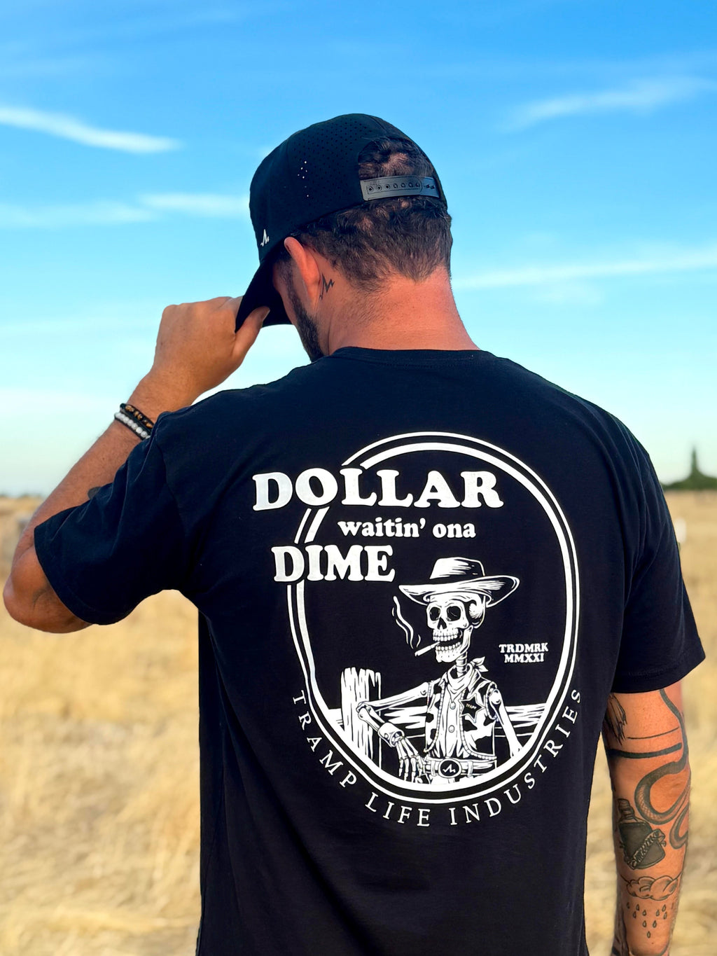man standing in a field wearing a black tramp life shirt with the words dollar waitin' ona dime and a skeleton smoking a cigarette leaning on a fence
