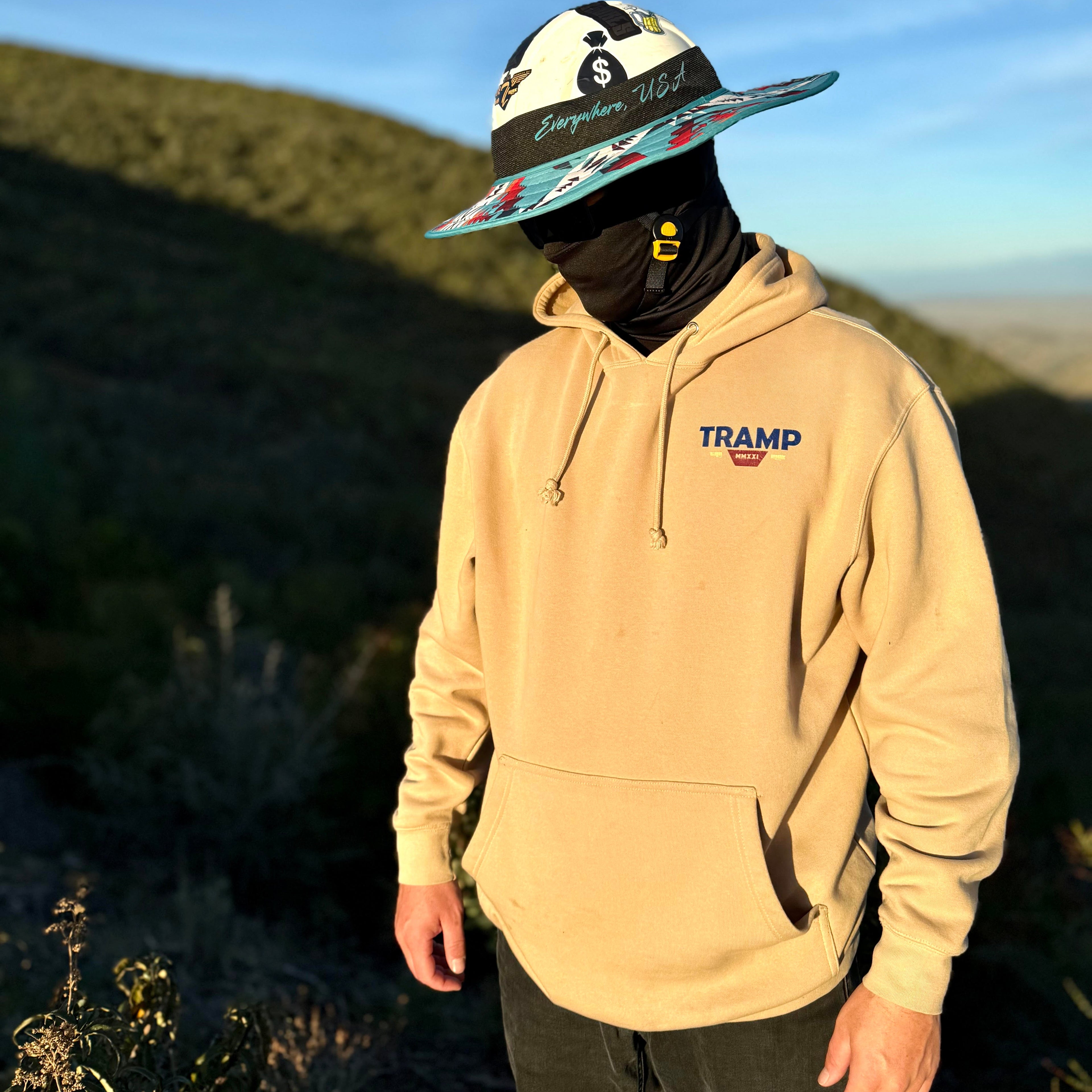 person wearing a tan hoodie from tramp life that says TRAMP on the front in blue writing. he is also wearing a turquoise fly shade and a black gaiter