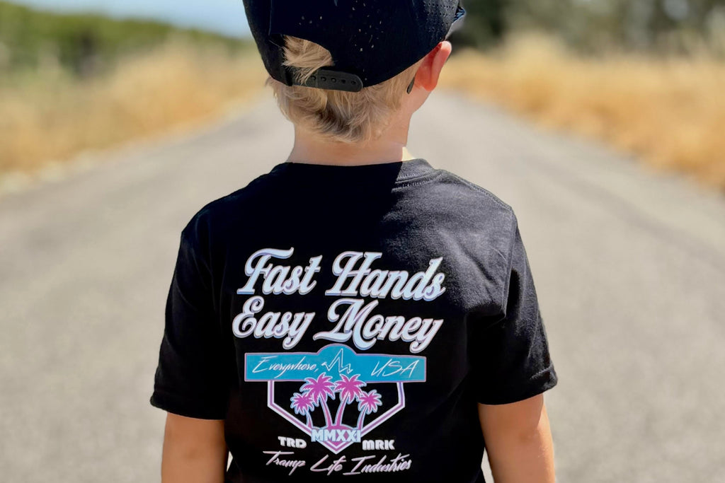 Child wearing a black t-shirt from tramp life with pink and blue palm tree and logos that says fast hands easy money, standing on a road.