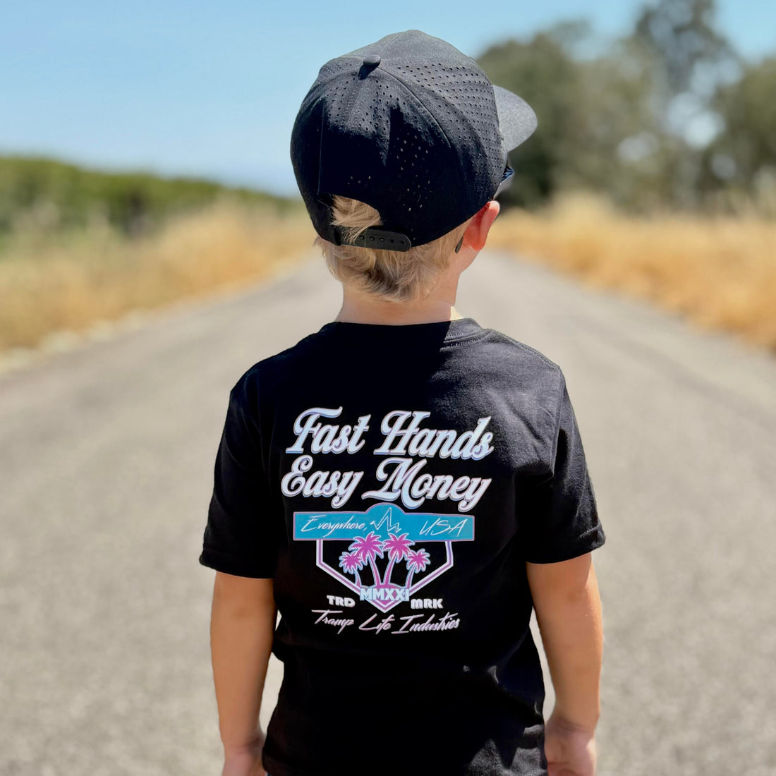 Child wearing a black t-shirt from tramp life with pink and blue palm tree and logos that says fast hands easy money, standing on a road.