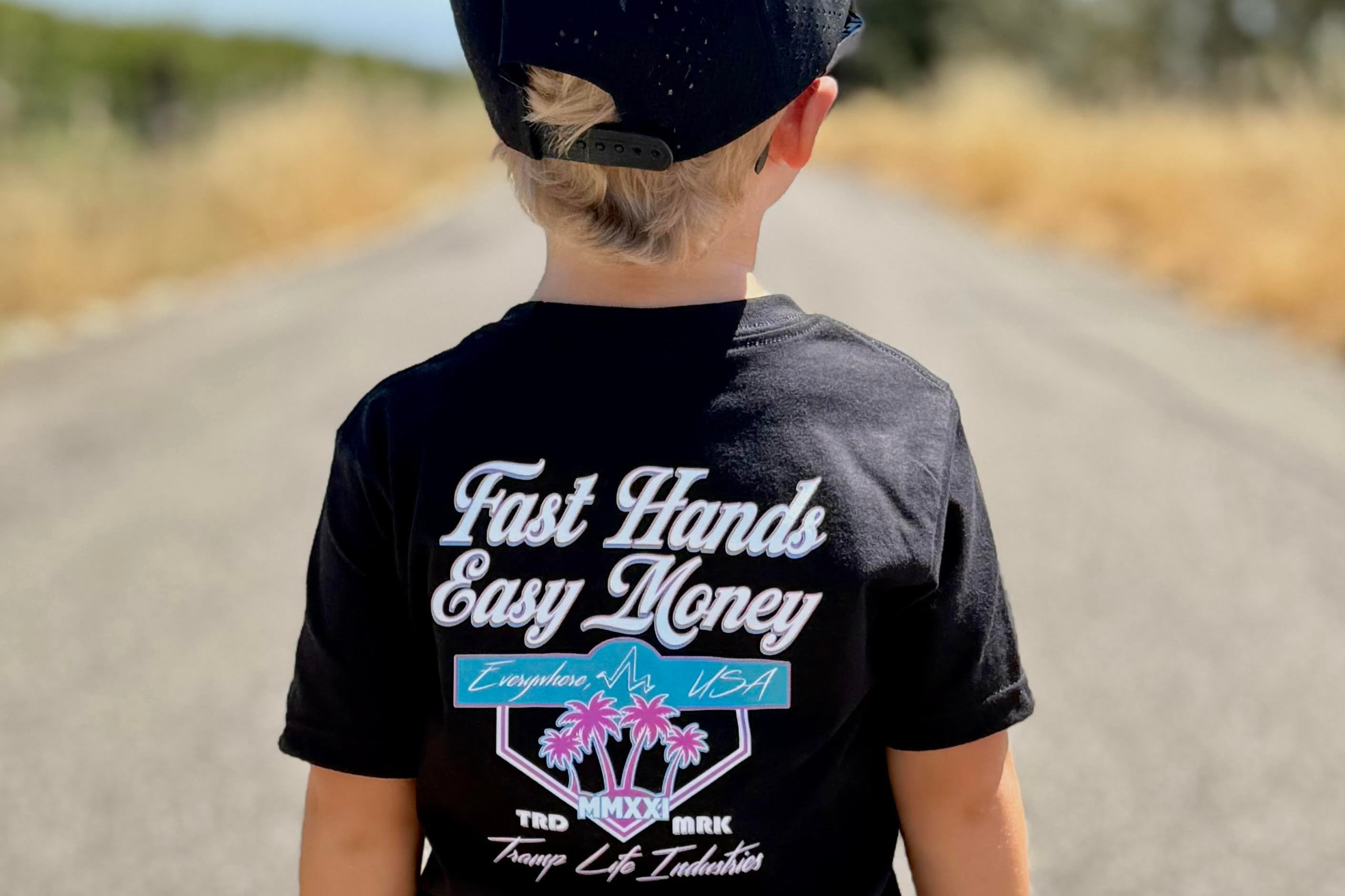 Child wearing a black t-shirt from tramp life with pink and blue palm tree and logos that says fast hands easy money, standing on a road.