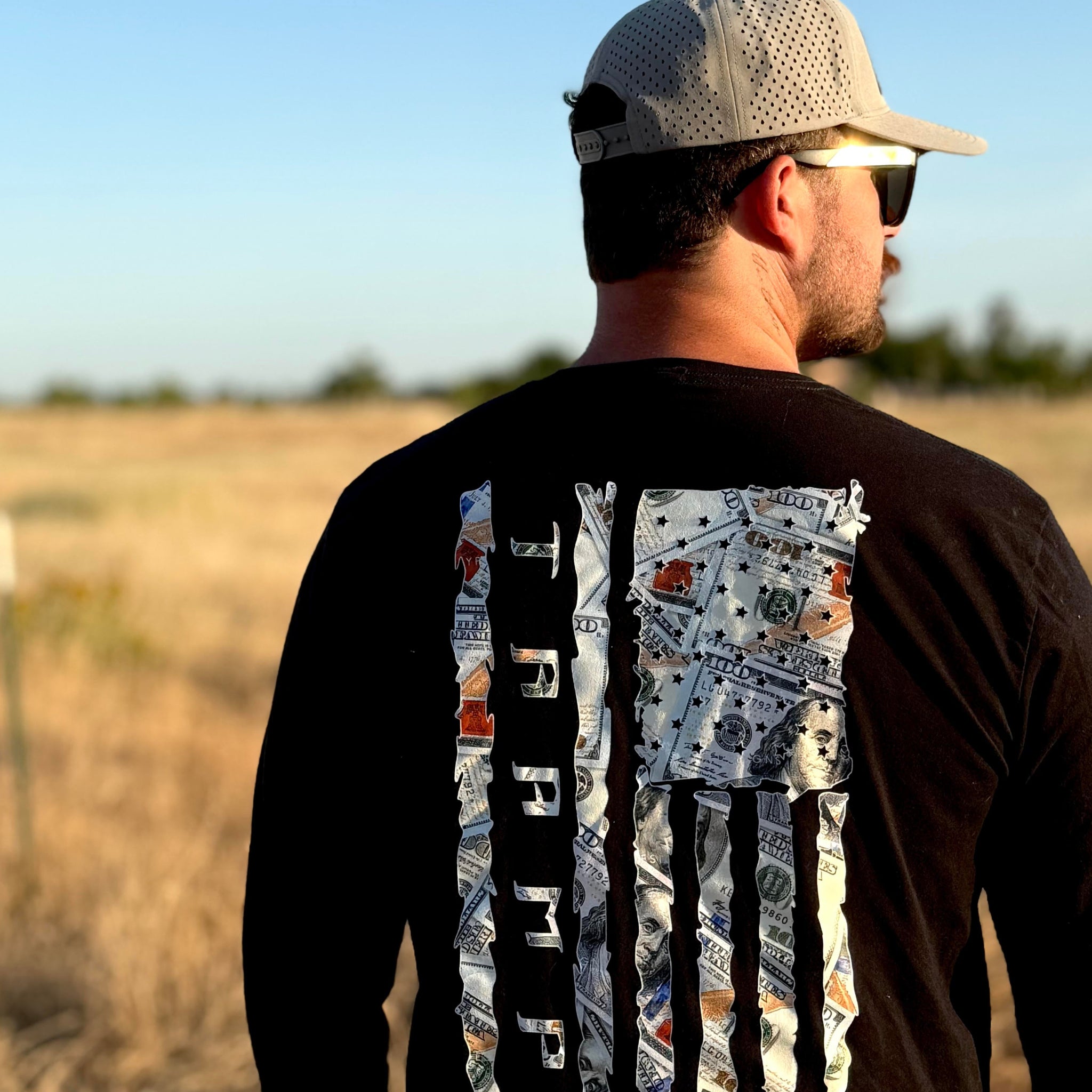 Man wearing a black long-sleeve shirt with a 100 dollar bills in a flag shape with the word tramp written down the middle standing in a field