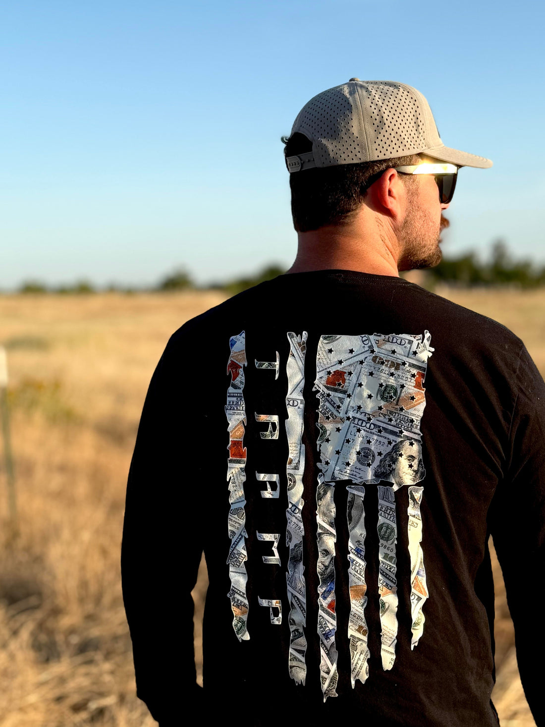 Man wearing a black long-sleeve shirt with a 100 dollar bills in a flag shape with the word tramp written down the middle standing in a field