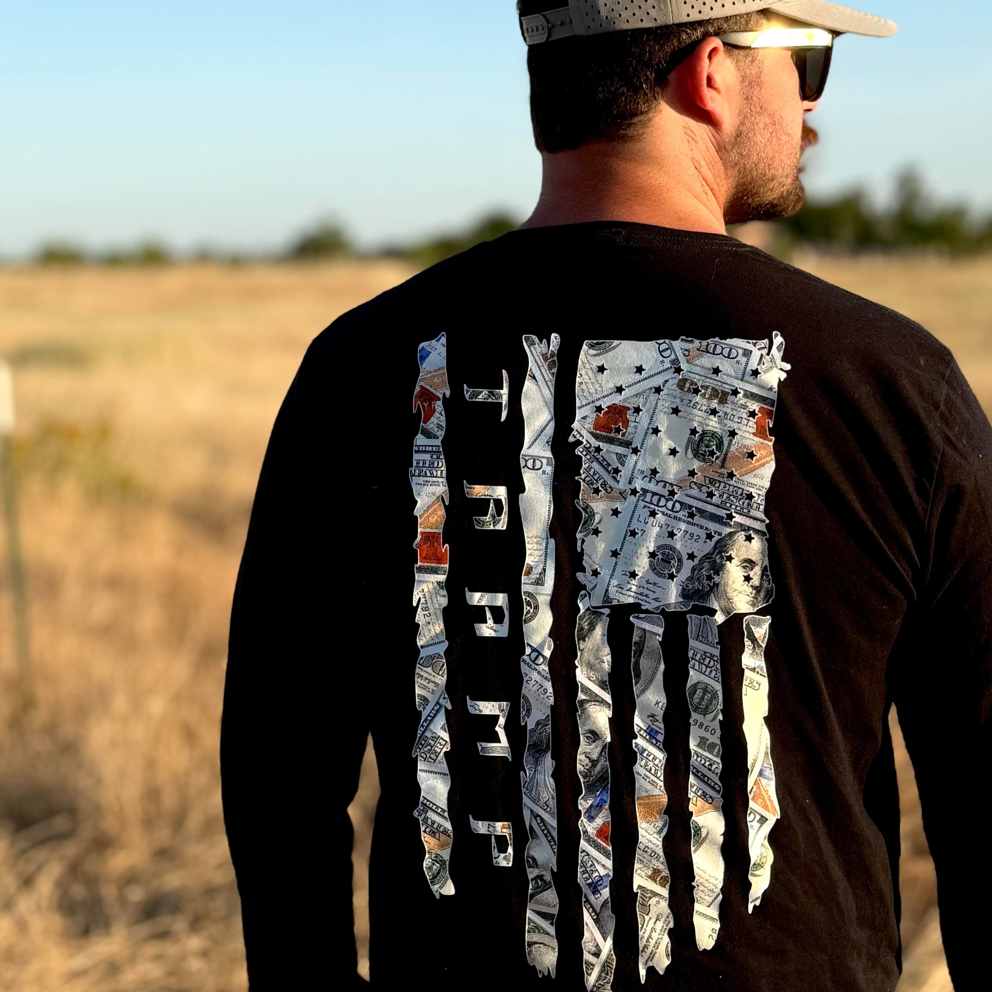 Man wearing a black long-sleeve shirt with a 100 dollar bills in a flag shape with the word tramp written down the middle standing in a field