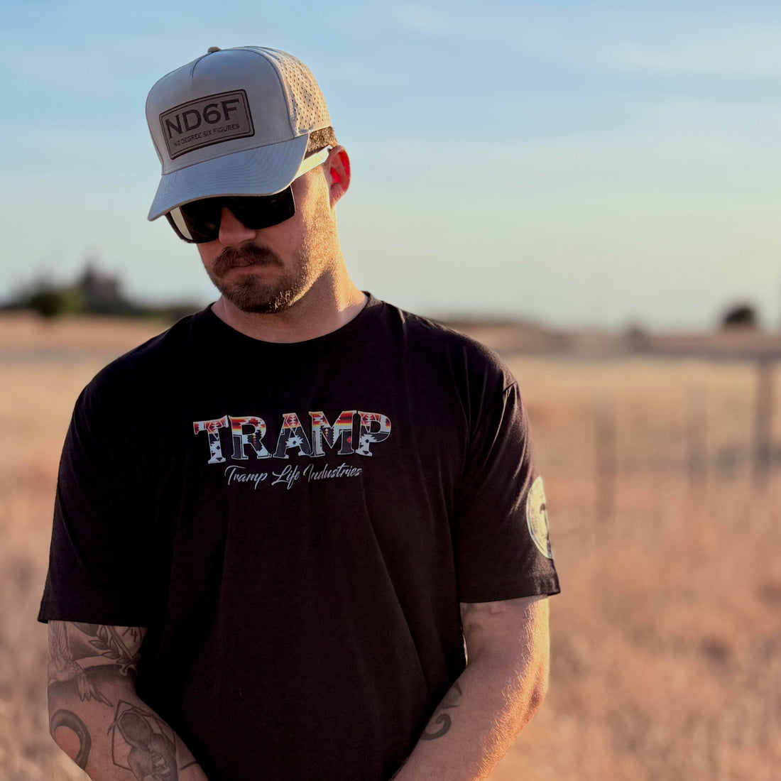 Person wearing a black 'TRAMP' t-shirt and cap in an open field.
