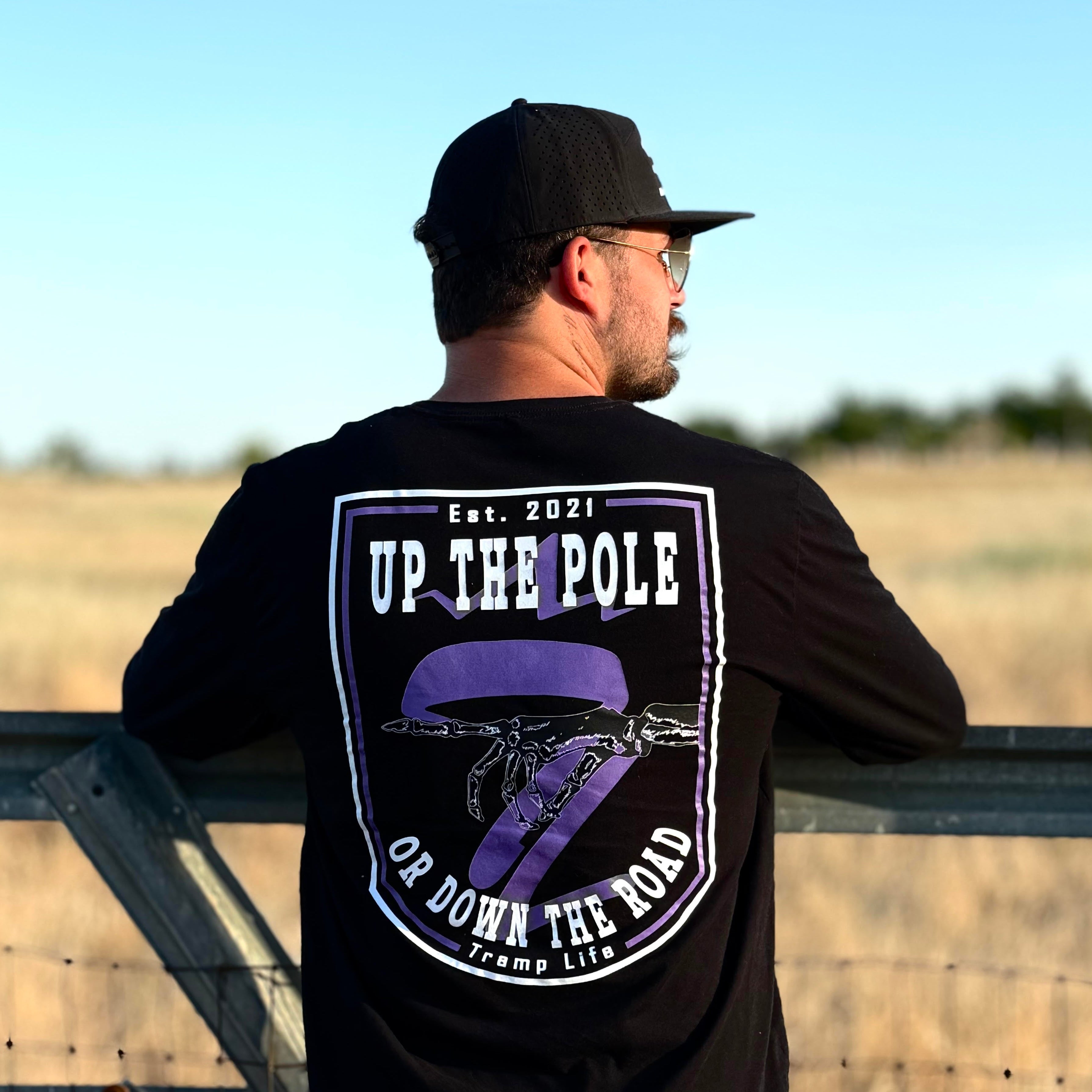 Man wearing a black t-shirt with a graphic design that says up the pole or down the road, standing by a fence in an open field.