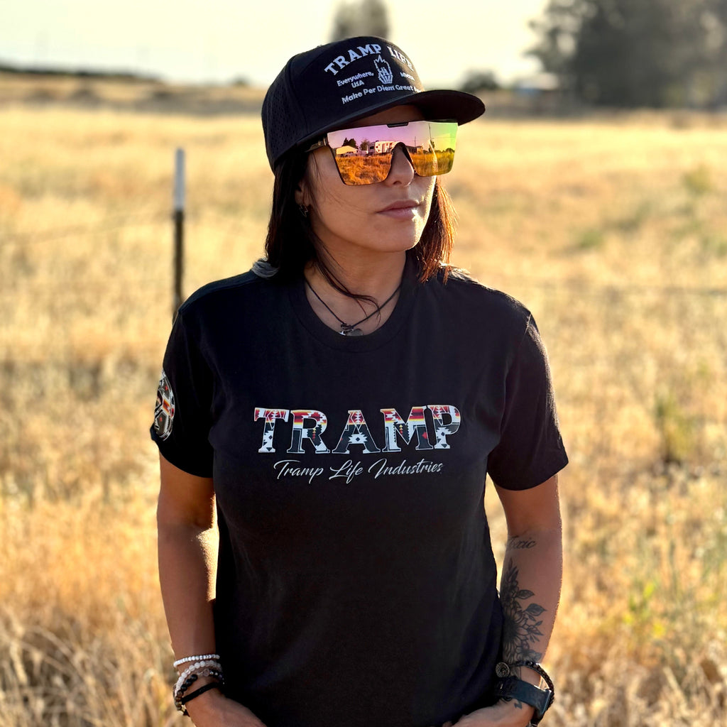 Person wearing a black 'TRAMP' t-shirt and cap in an open field.