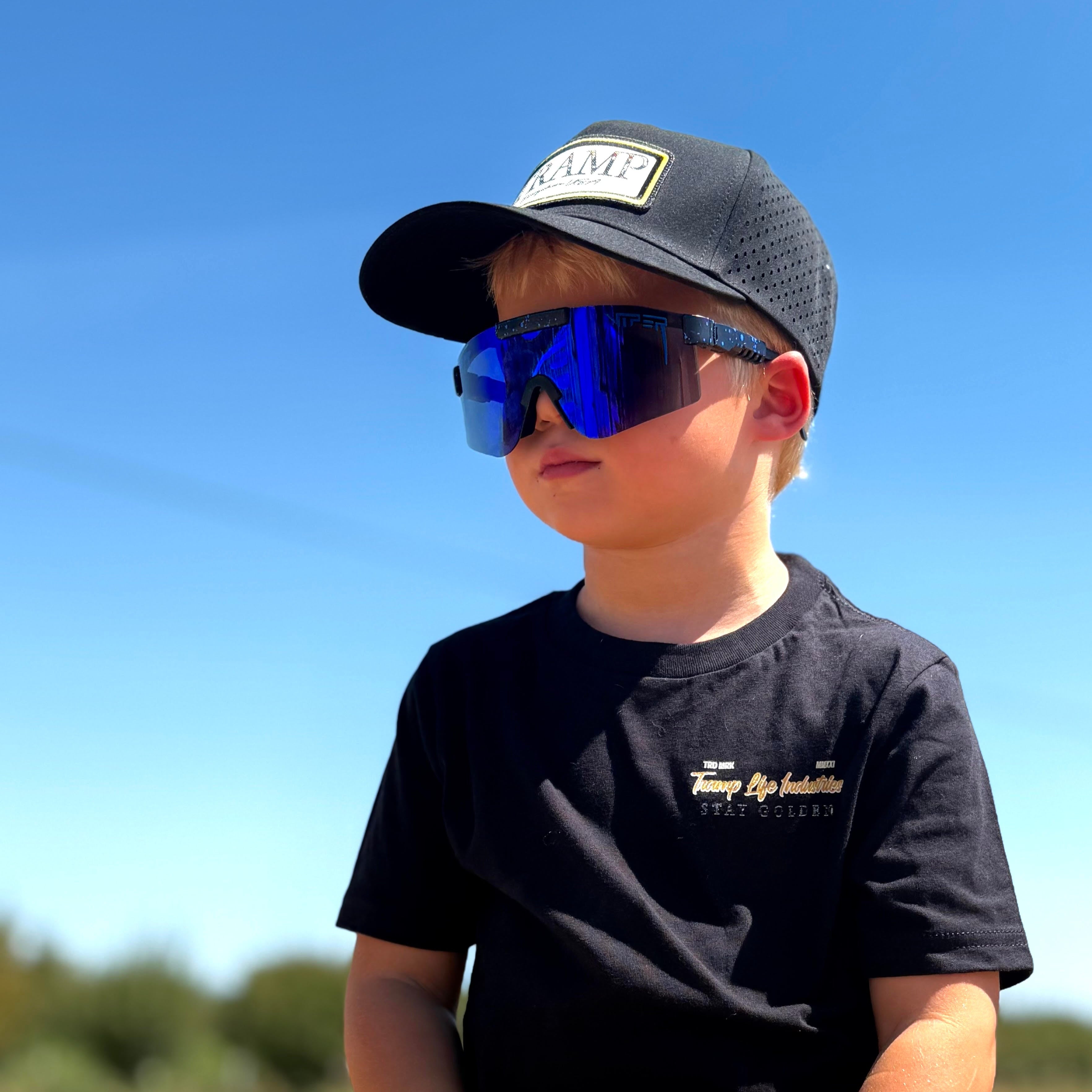 Child wearing a black cap and sunglasses sitting in a field with a clear blue sky.