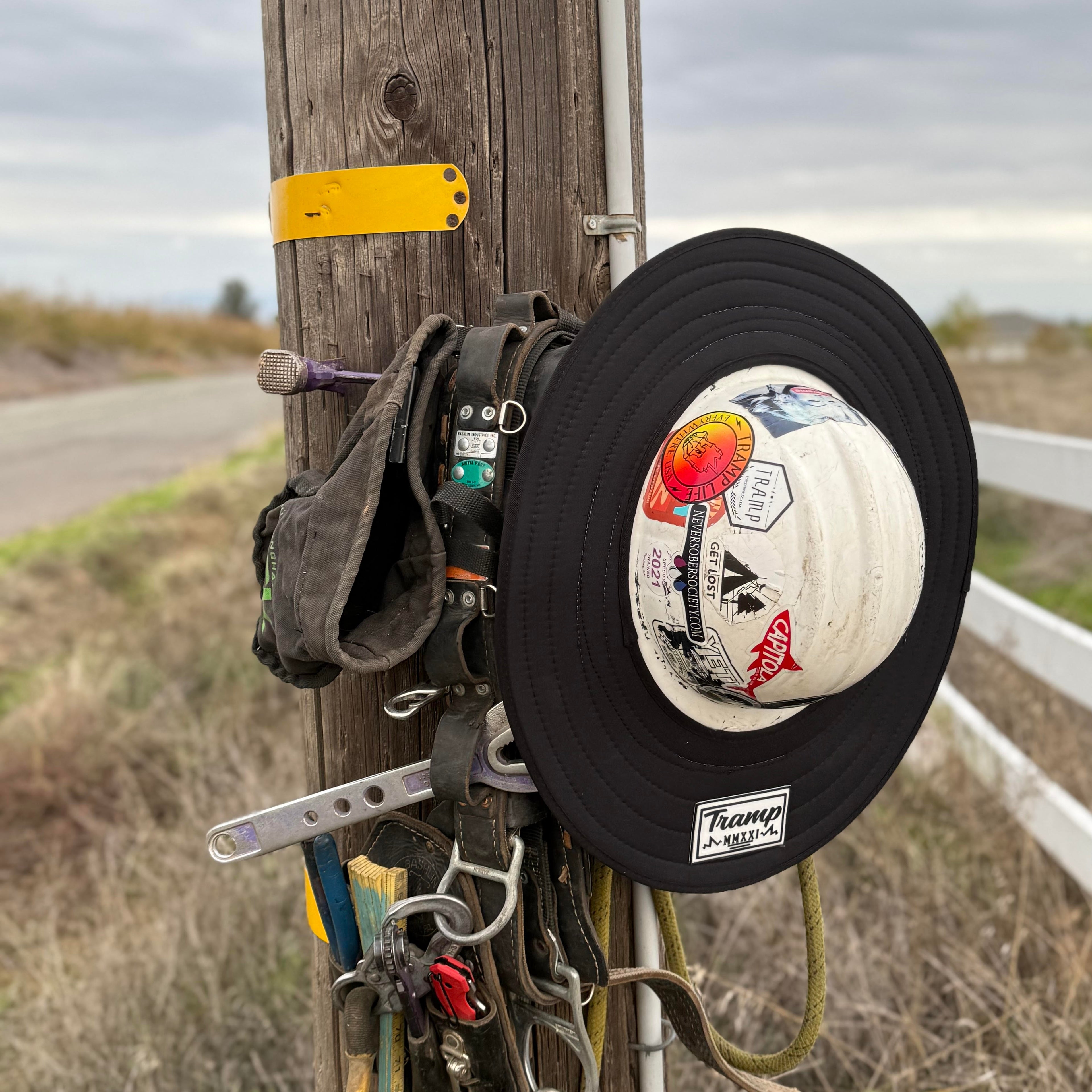 A black print hard hat sunshade from tramp life industries. Features a completely black print and attaches to full brim hard hats with an elastic band. the hardhat and sunshade are on a power pole hanging next to lineman climbing gear.