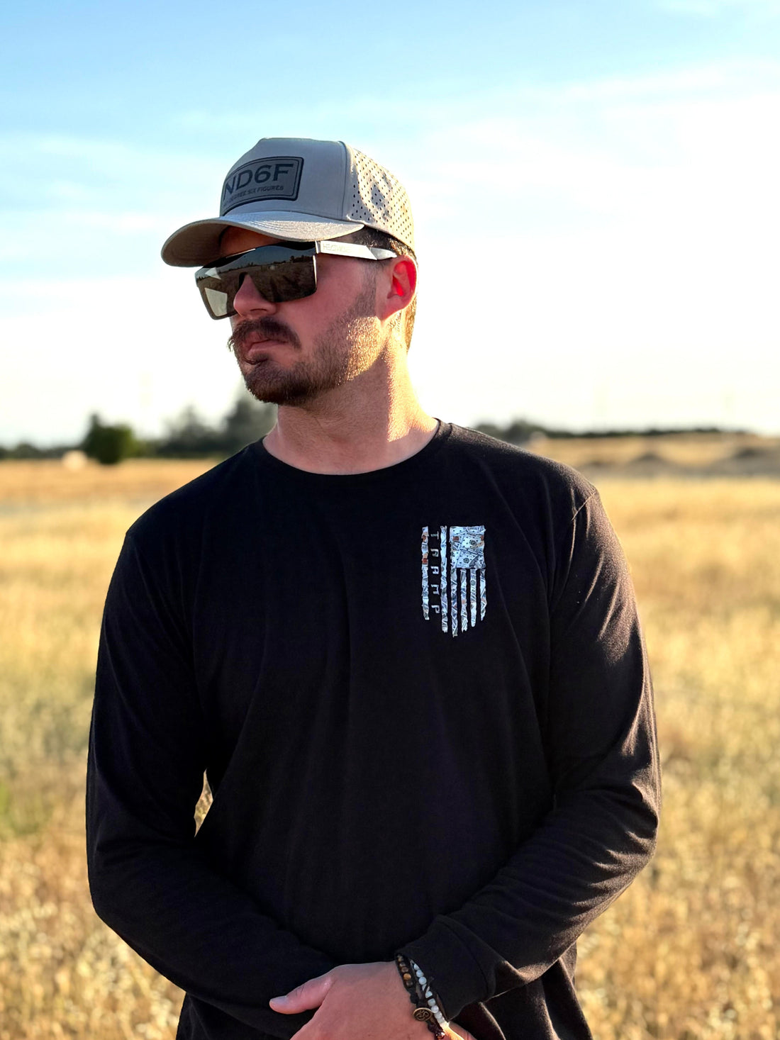 Man wearing a black long-sleeve shirt with a 100 dollar bills in a flag shape with the word tramp written down the middle standing in a field