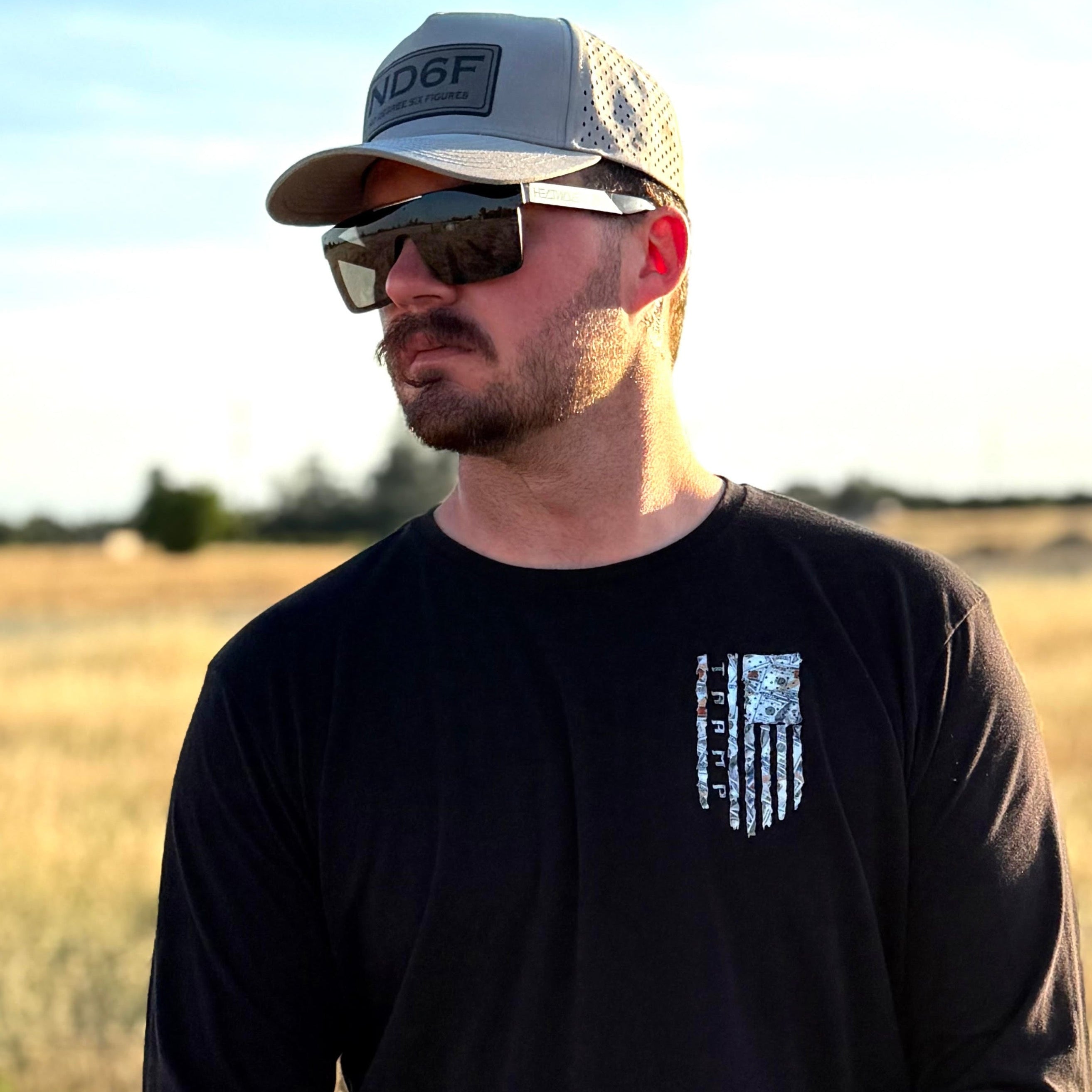 Man standing in field with a flag made from 100 dollar bills and the word tramp inside of it, standing in a field with a gray tramp life hat on