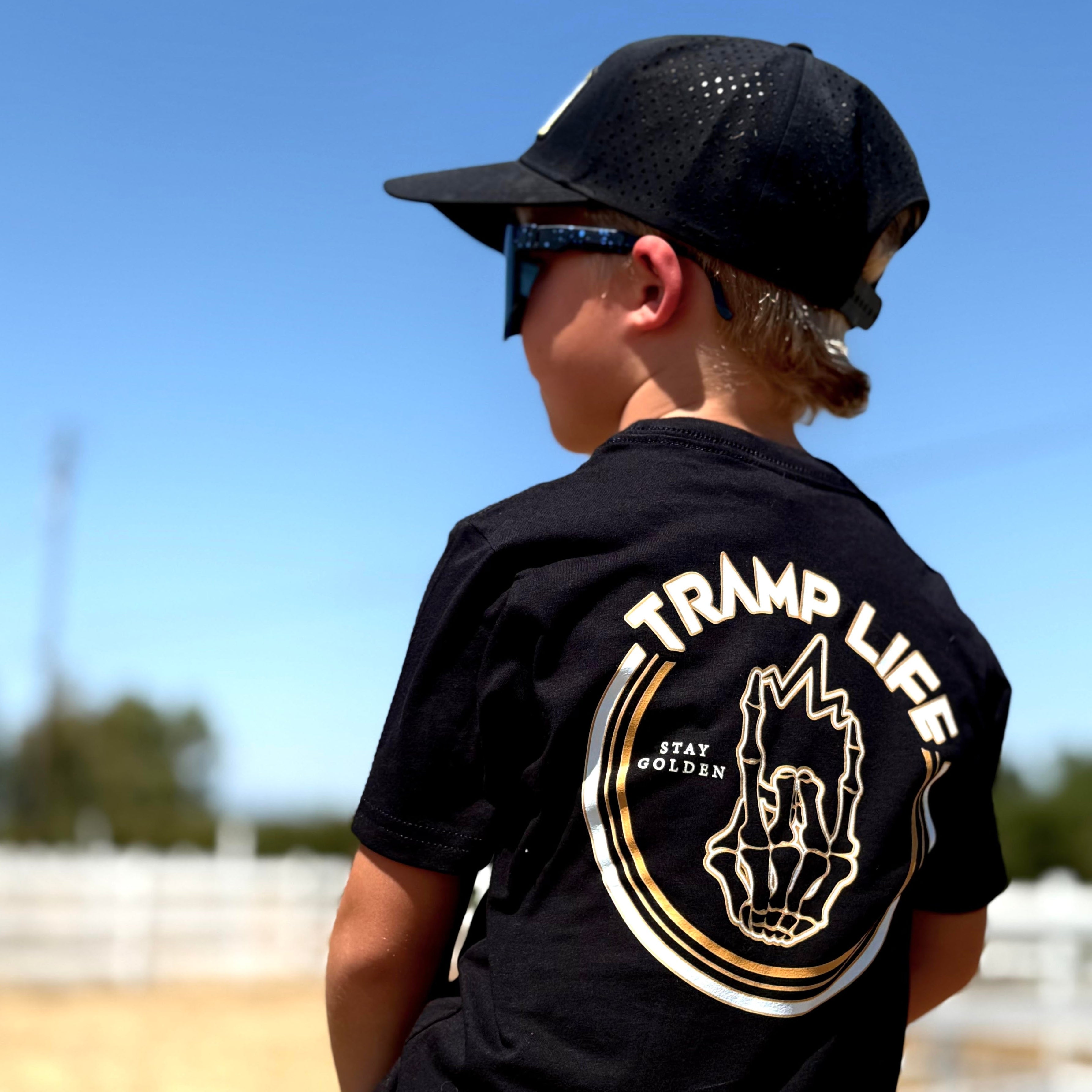 Child wearing a black 'Tramp Life' t-shirt with a gold hand graphic outdoors.