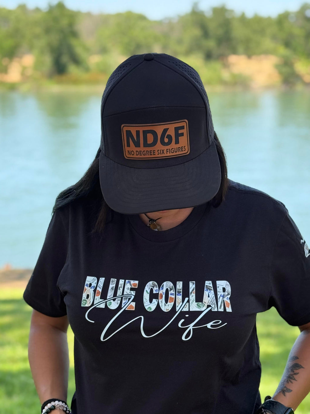 Person wearing a black t-shirt with 'Blue Collar Wife' text and a cap with 'ND6F' logo outdoors by a lake.