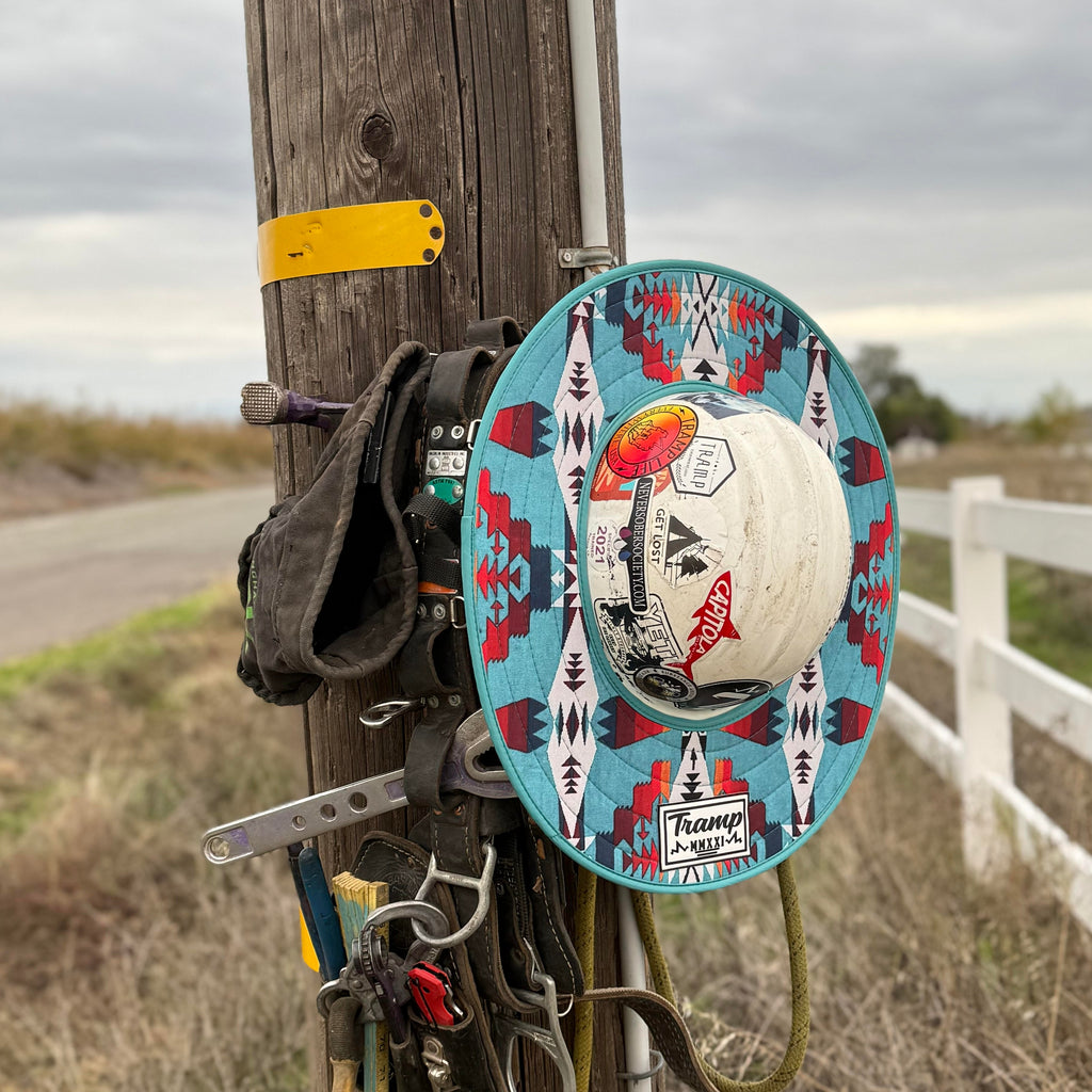 A western turquoise print hard hat sunshade from tramp life industries. Features a western turquoise print and attaches to full brim hard hats with an elastic band. the hardhat and sunshade are on a power pole hanging next to lineman climbing gear.