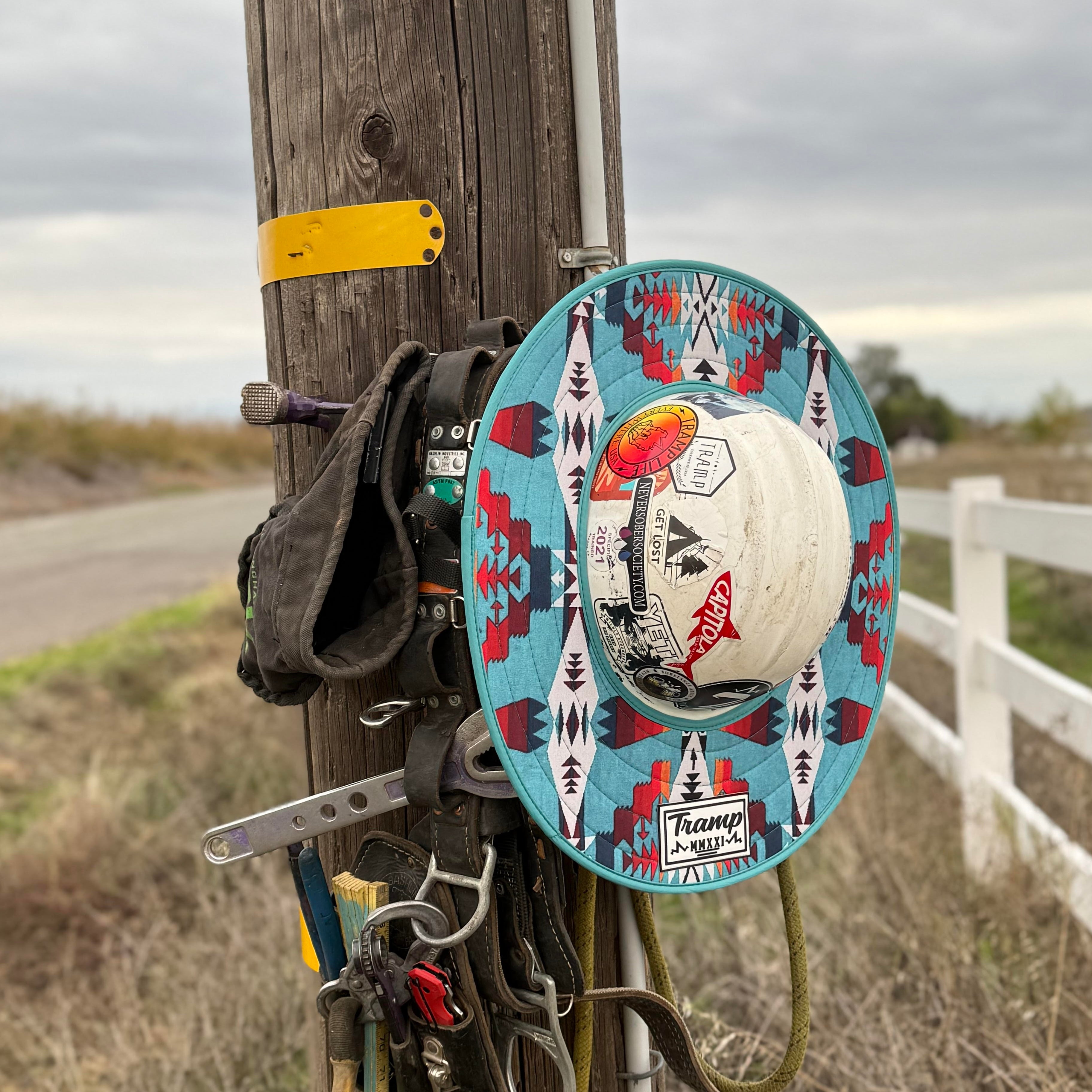 A western turquoise print hard hat sunshade from tramp life industries. Features a western turquoise print and attaches to full brim hard hats with an elastic band. the hardhat and sunshade are on a power pole hanging next to lineman climbing gear.