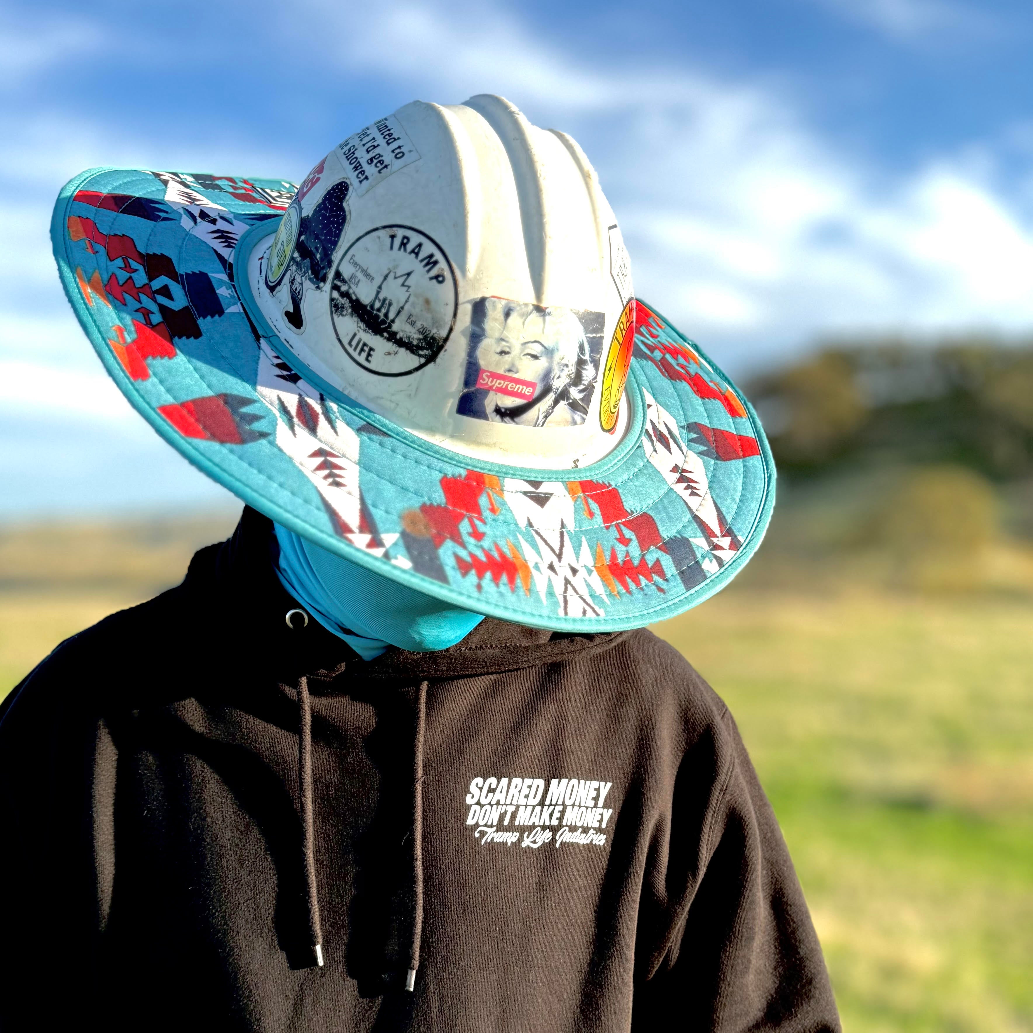 man standing in a field wearing a hard hat with the turquoise sunshade from tramp life. has a black hoodie on that says scared money dont make money