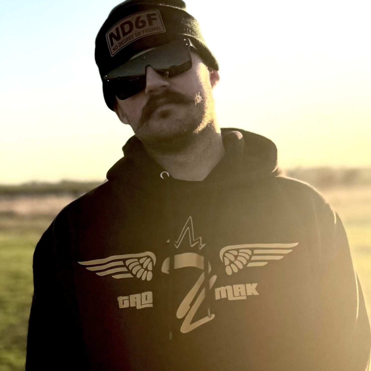 a man wearing a black hoodie from tramp life industries that has a gold TL with wings on the chest. he is also wearing a beanie that says ND6F and no degree six figures