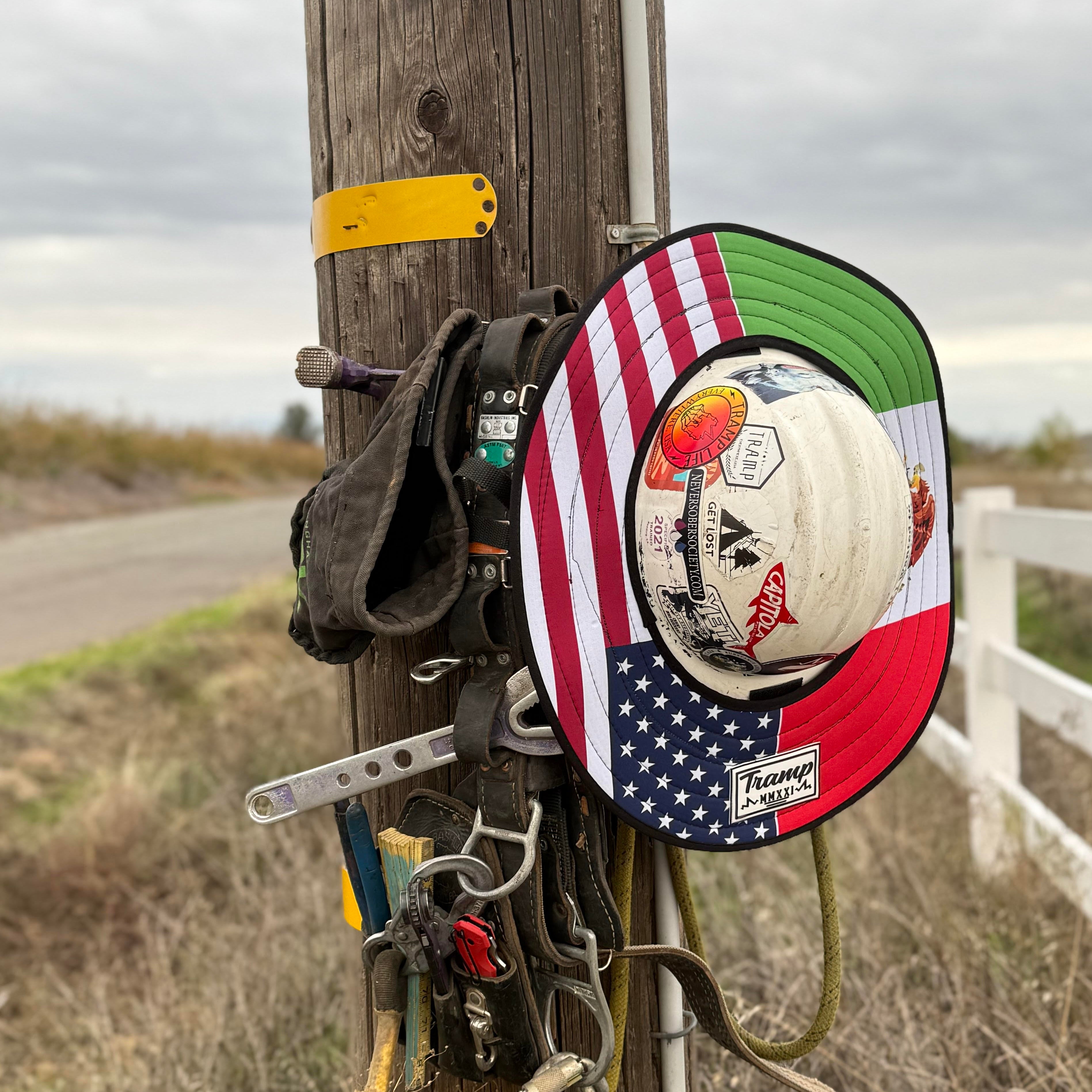 A half mexican flag half ameican flag print hard hat sunshade from tramp life industries. Features a half american flag and half mexican flag and attaches to full brim hard hats with an elastic band. the hardhat and sunshade are on a power pole hanging next to lineman climbing gear.