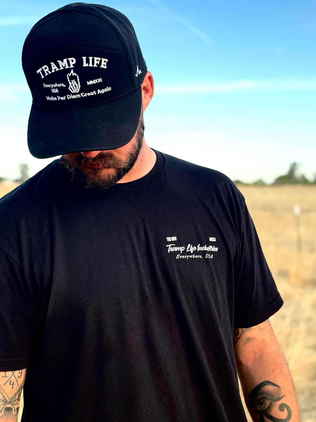 man standing in a field with a black tramp life shirt on that says tramp life industries and a black hat that says make per diem great again